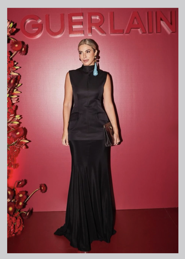 Woman in a black sleeveless gown holding a clutch, standing in front of a pink backdrop with red flowers to the left and the word 'GUERLAIN' at the top.
