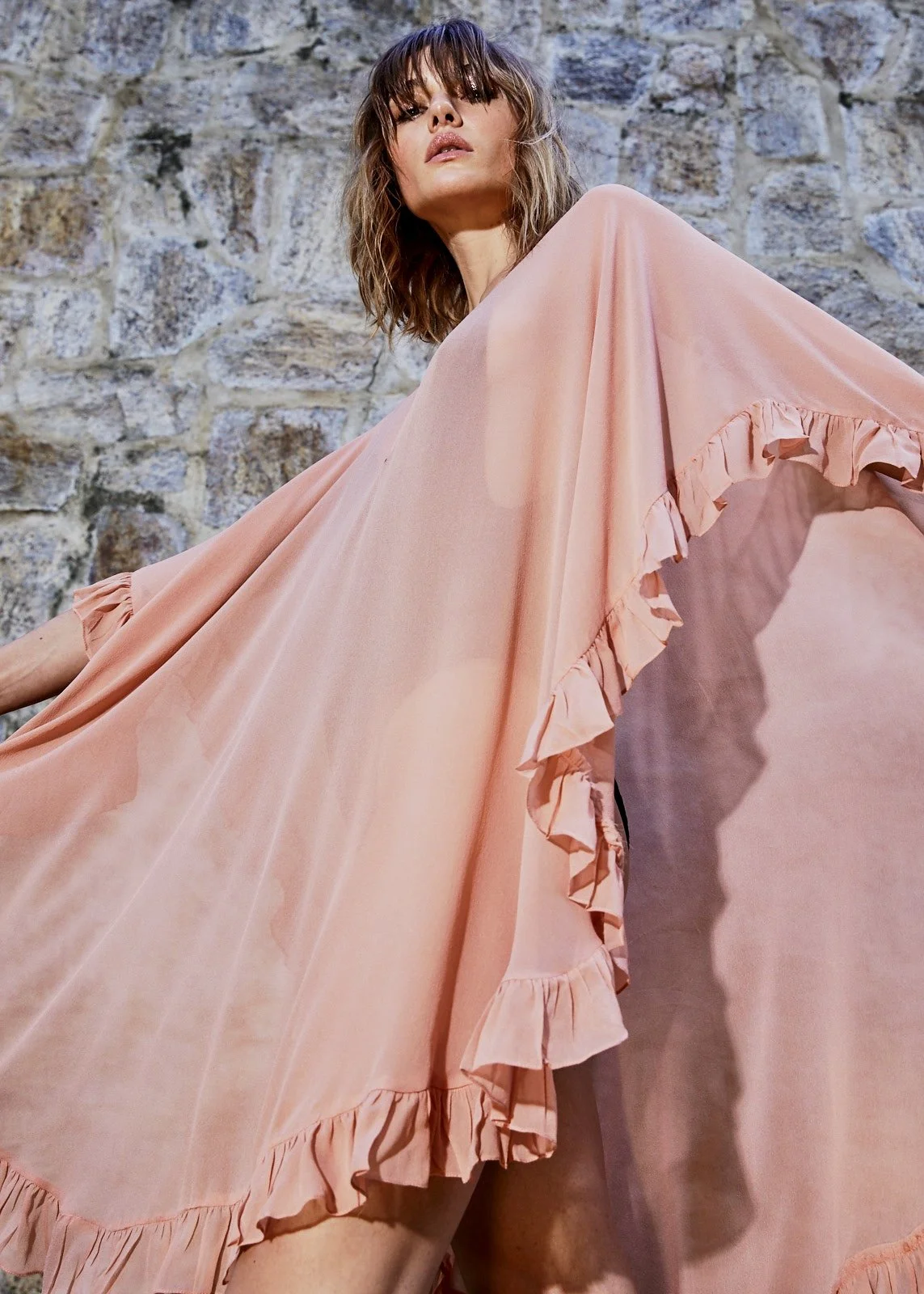 A woman with shoulder-length wavy hair posing against a stone wall, wearing a flowing peach-colored dress with ruffled edges.