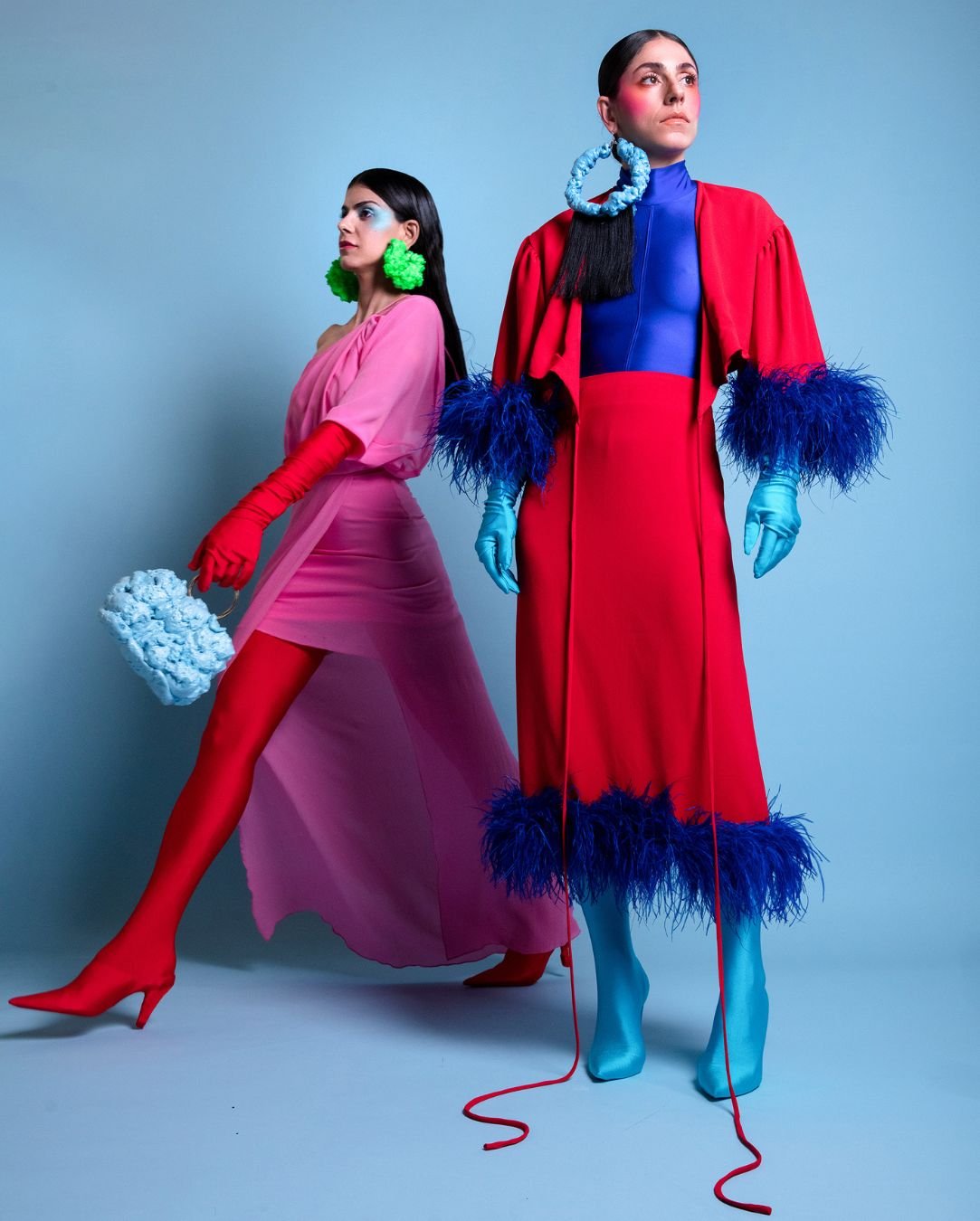 Two women in bold, colorful fashion with exaggerated accessories and footwear, standing against a light blue background.