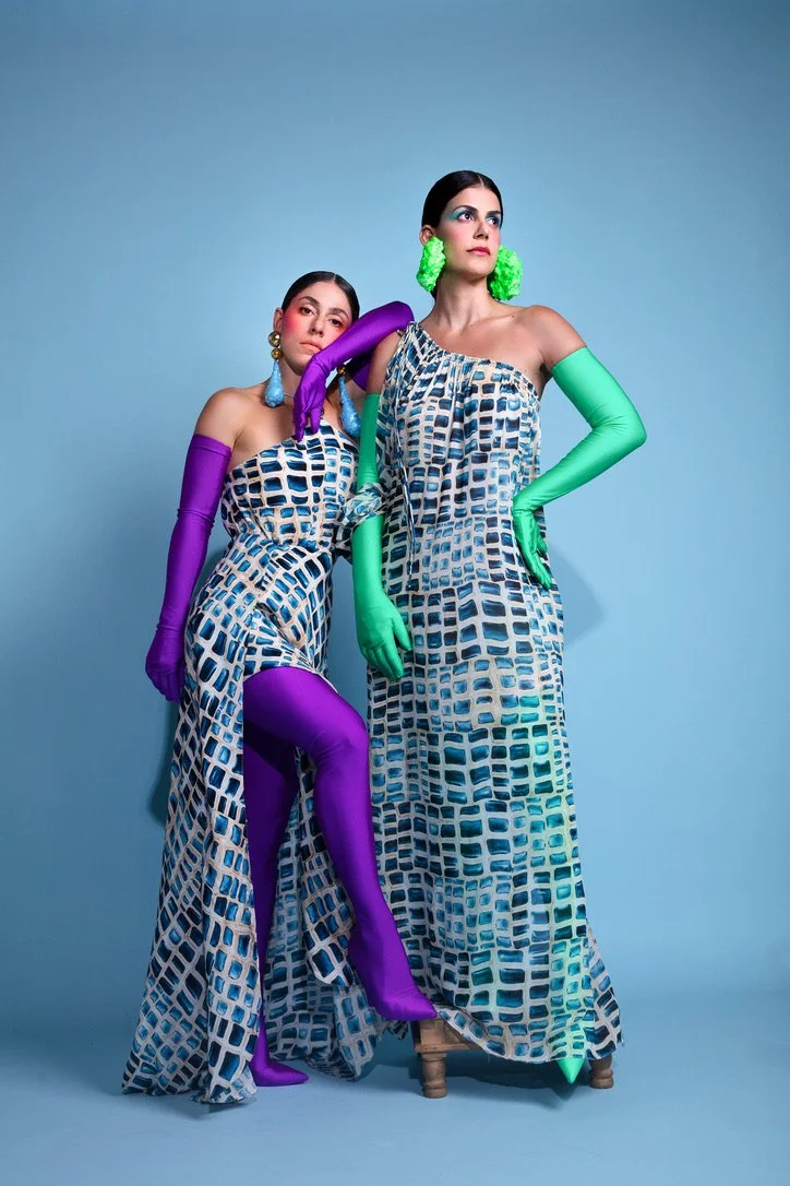 Two women pose in matching blue patterned dresses with large grid-like designs against a light blue background. They wear colorful long gloves, earrings, and green and purple accessories, with bold makeup, creating a fashion-forward, artistic look.
