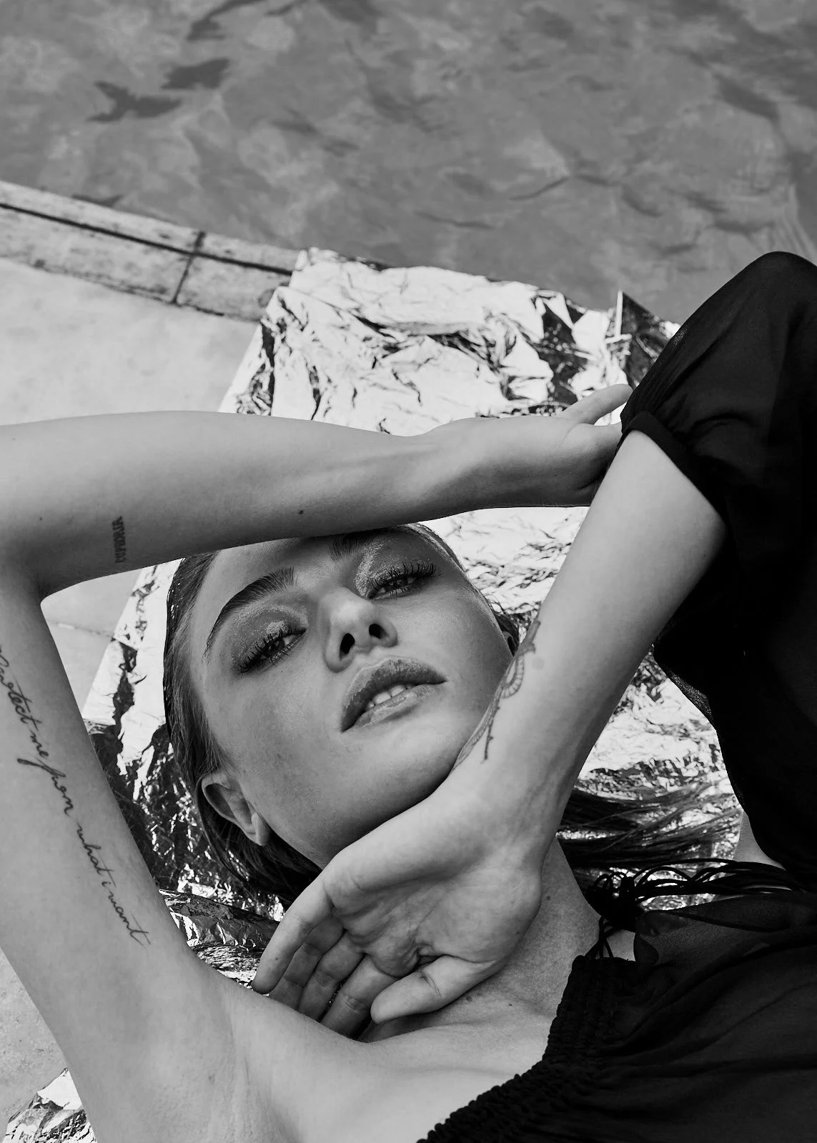 Black and white photograph of a woman with tattoos, lying on a reflective surface, gazing at the camera with her arms above her head, in front of a body of water.