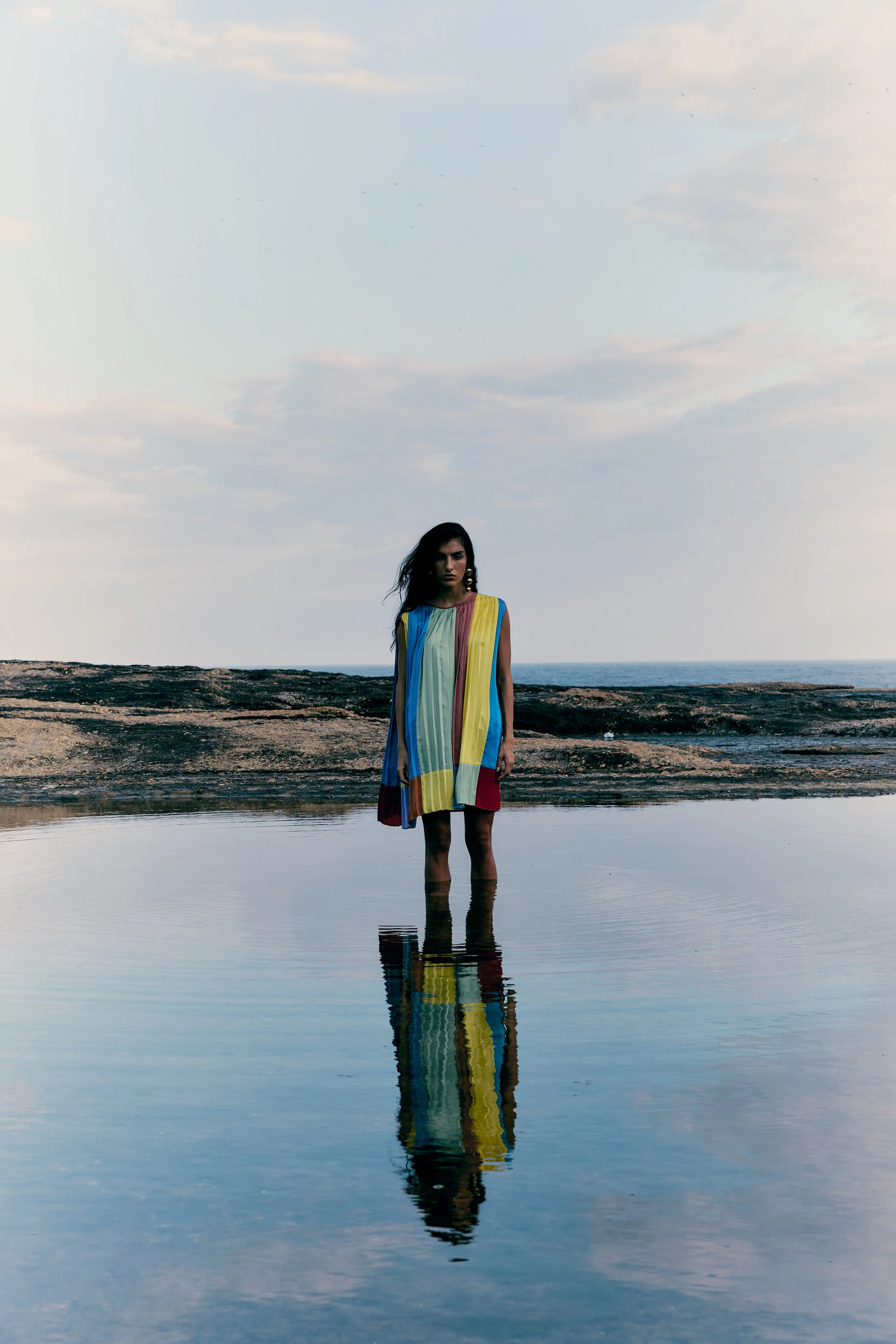 A woman standing in shallow water at the beach, wearing a colorful striped dress, with a cloudy sky and ocean in the background, and her reflection in the water.