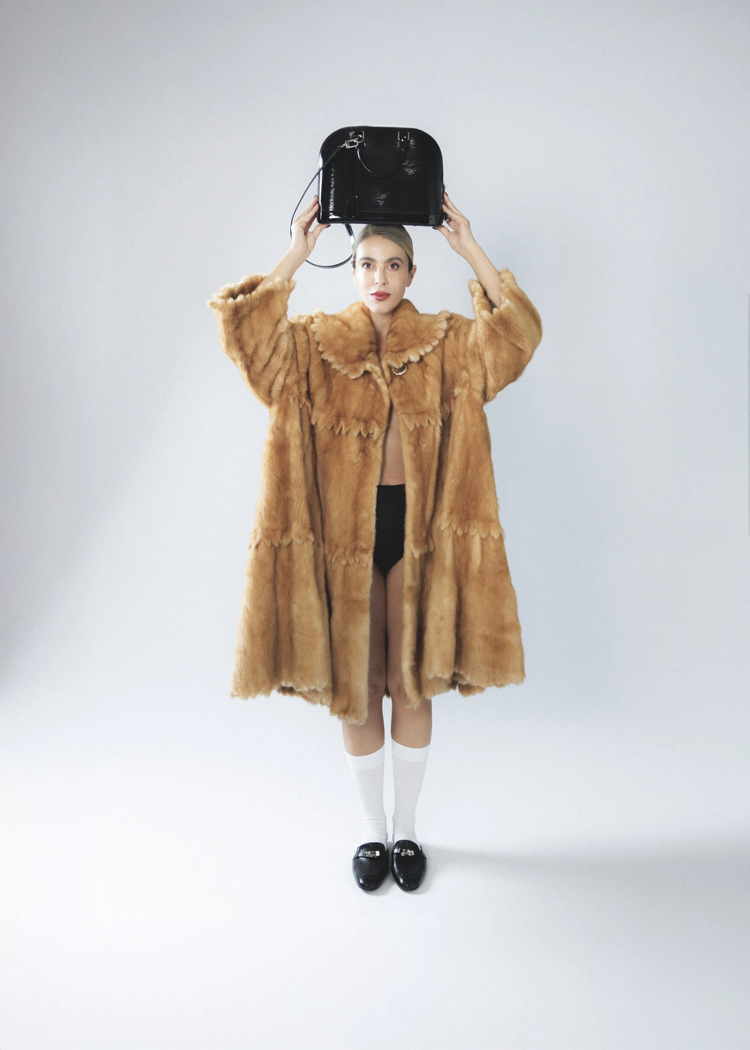 A woman in a fur coat holding a black handbag above her head, wearing white knee-high socks and black shoes, standing against a plain light grey background.