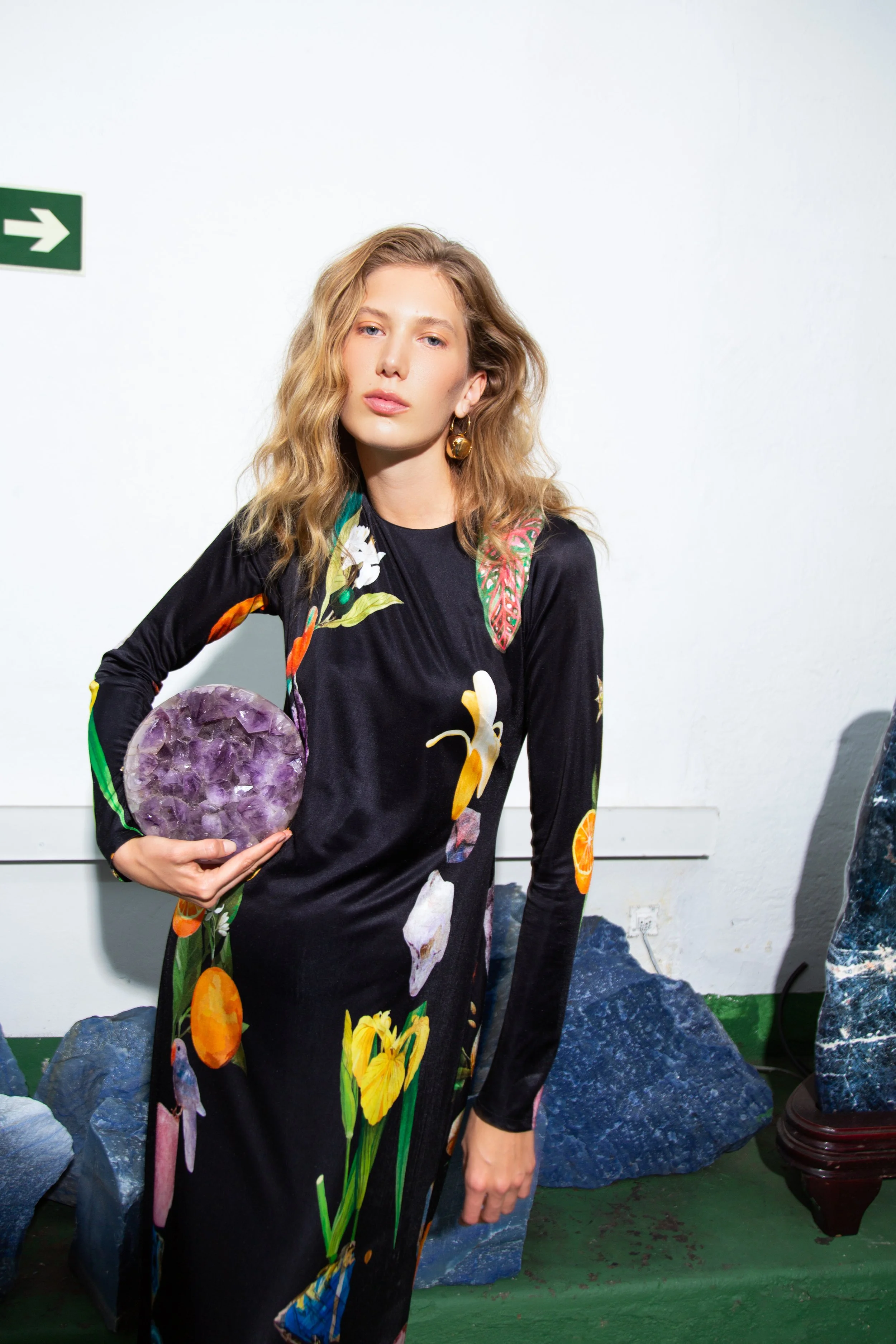 A woman with long, wavy blonde hair wearing a colorful floral dress holding a purple crystal geode, standing in front of a white wall with rocks on the floor.