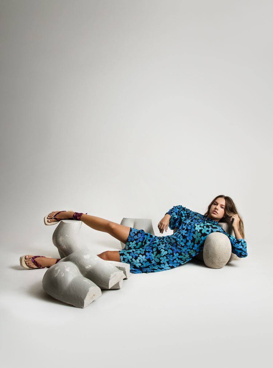 A woman in a blue floral dress lying on the floor, surrounded by broken concrete pieces, with one arm resting on a stone and one leg extended onto a concrete block, against a plain white background.