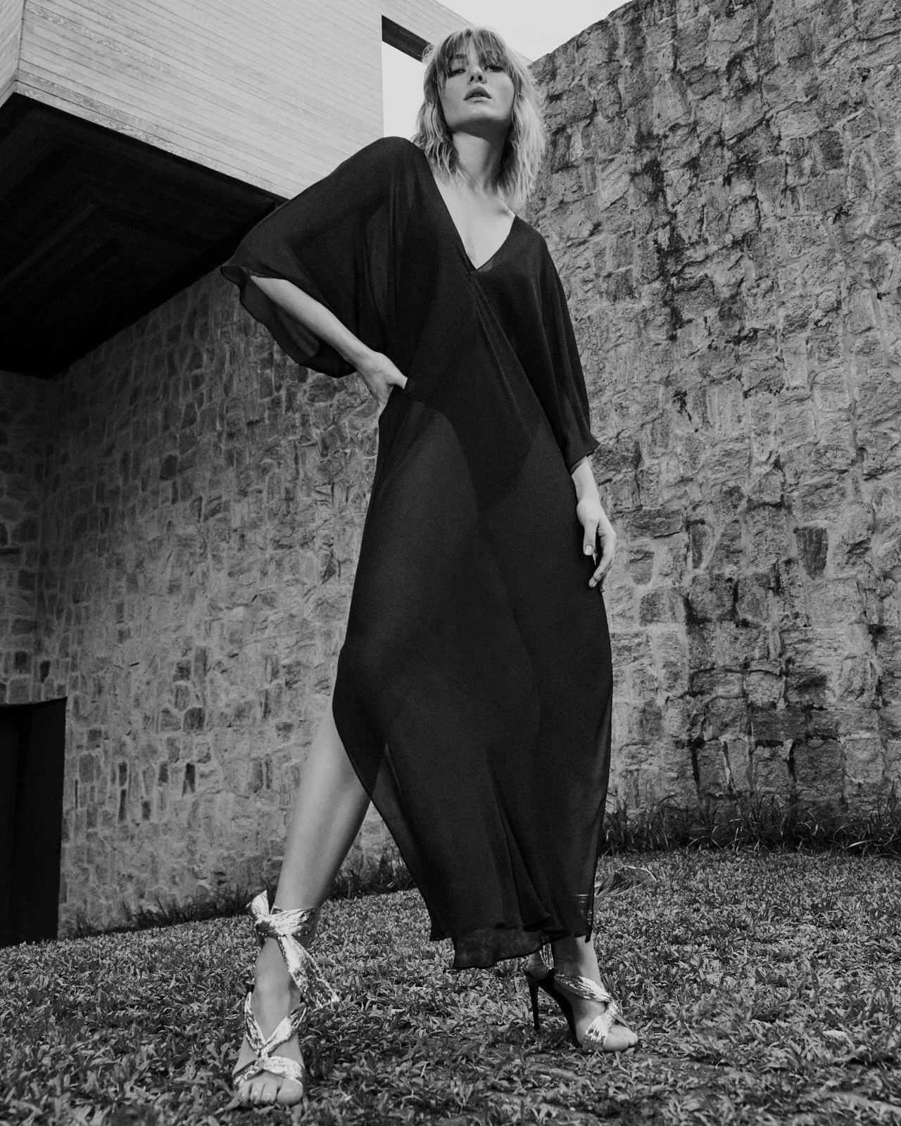 Black and white photo of a woman wearing a long, sheer dress and high heels, standing outdoors against a stone wall, with a lawn and some plants in the background.