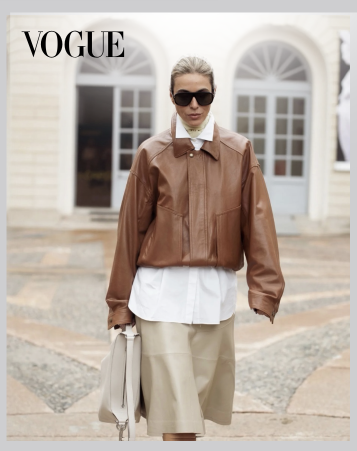 A woman walking outdoors, wearing large black sunglasses, a brown leather jacket, a white shirt, and beige pants, holding a light-colored handbag.
