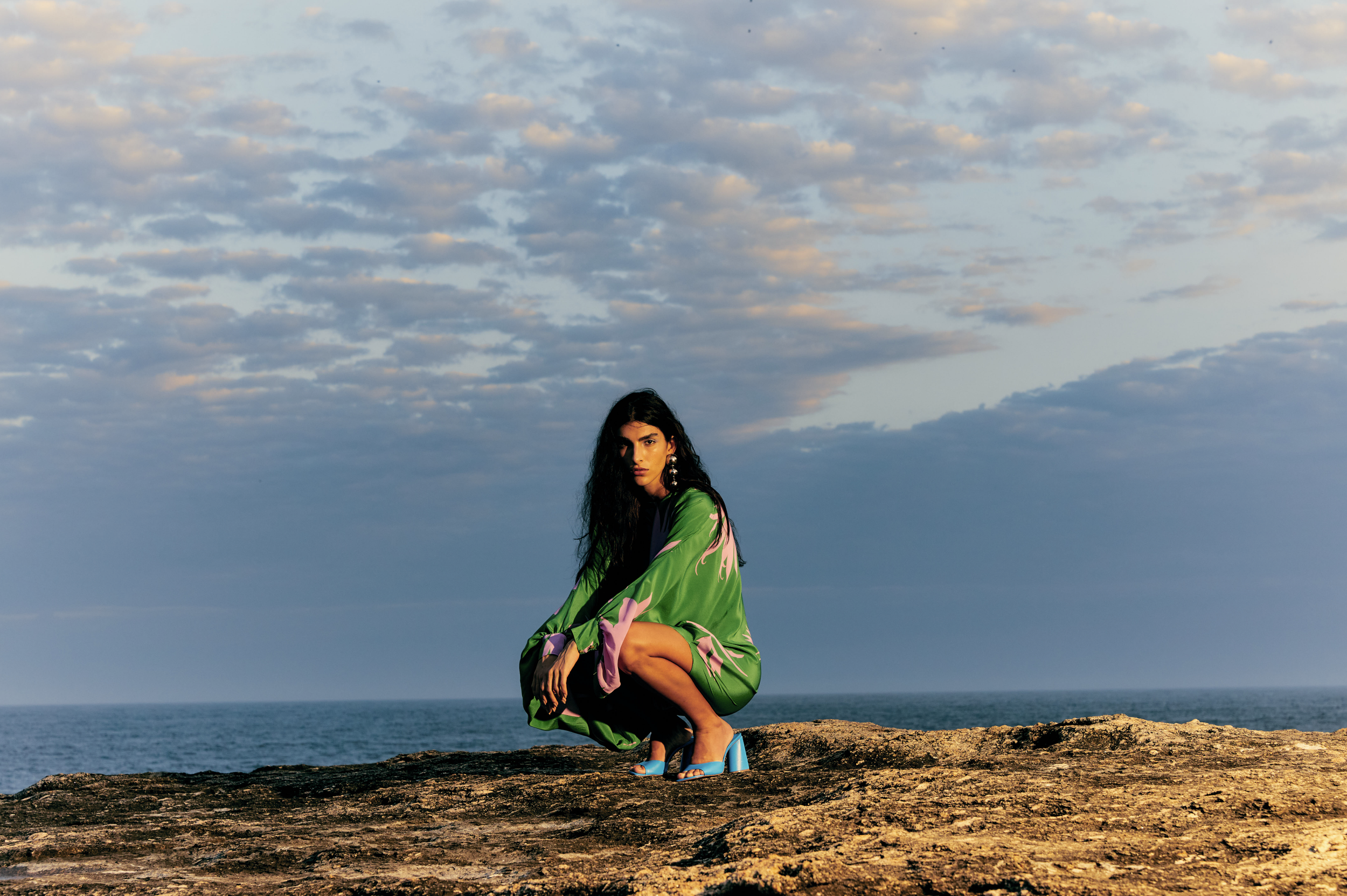A woman with long dark hair squatting on a rocky surface near the ocean, wearing a green and pink patterned dress with open-toe blue high-heeled shoes, against a backdrop of a partly cloudy sky.