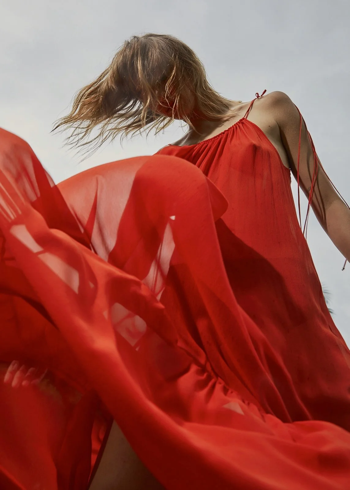 A woman wearing a flowing red dress with thin straps, standing outdoors under a cloudy sky, with her head tilted downward and her hair blowing in the wind.