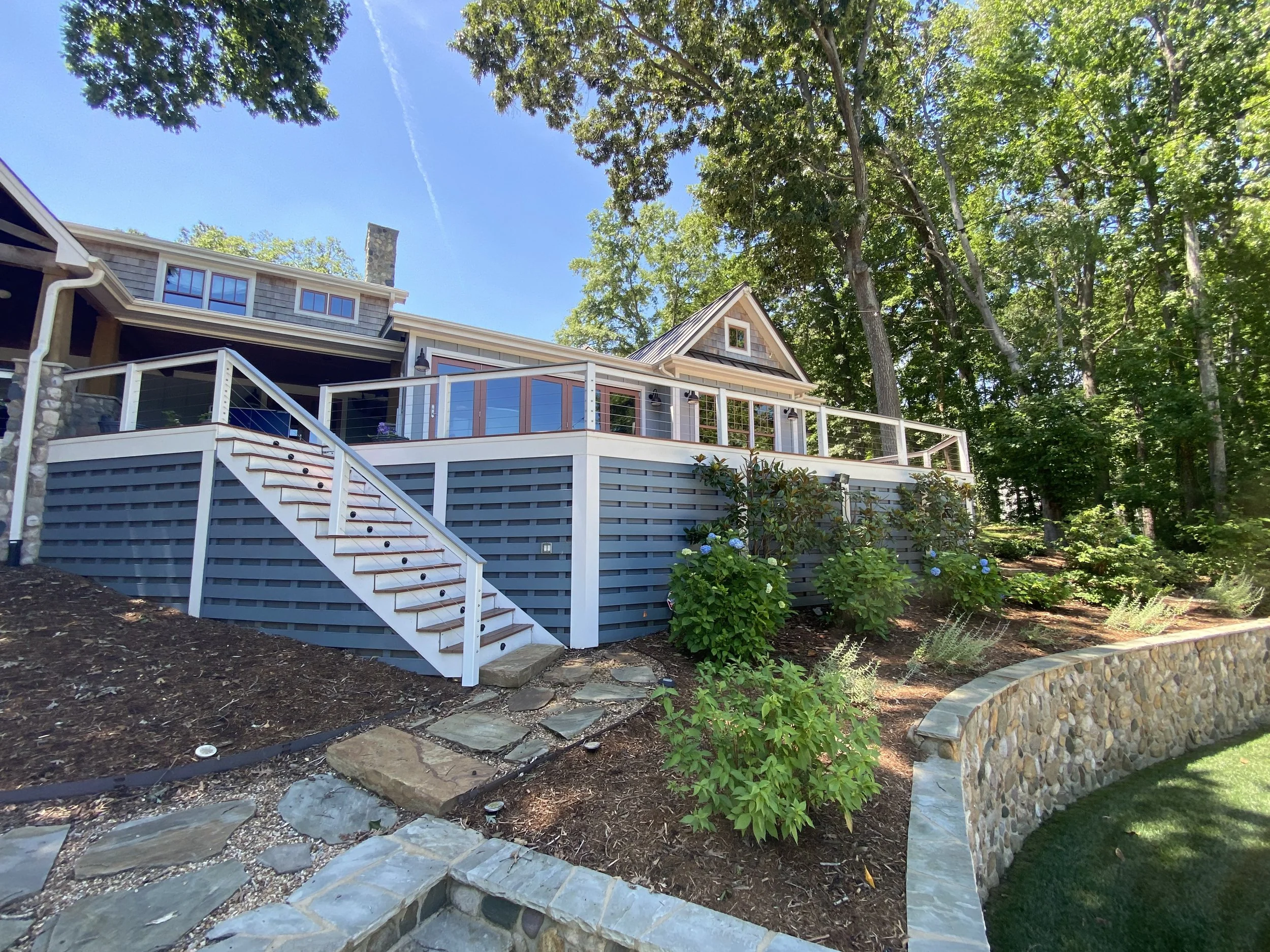 A house with a deck, surrounded by trees and a landscaped yard with stone accents and flowering bushes.
