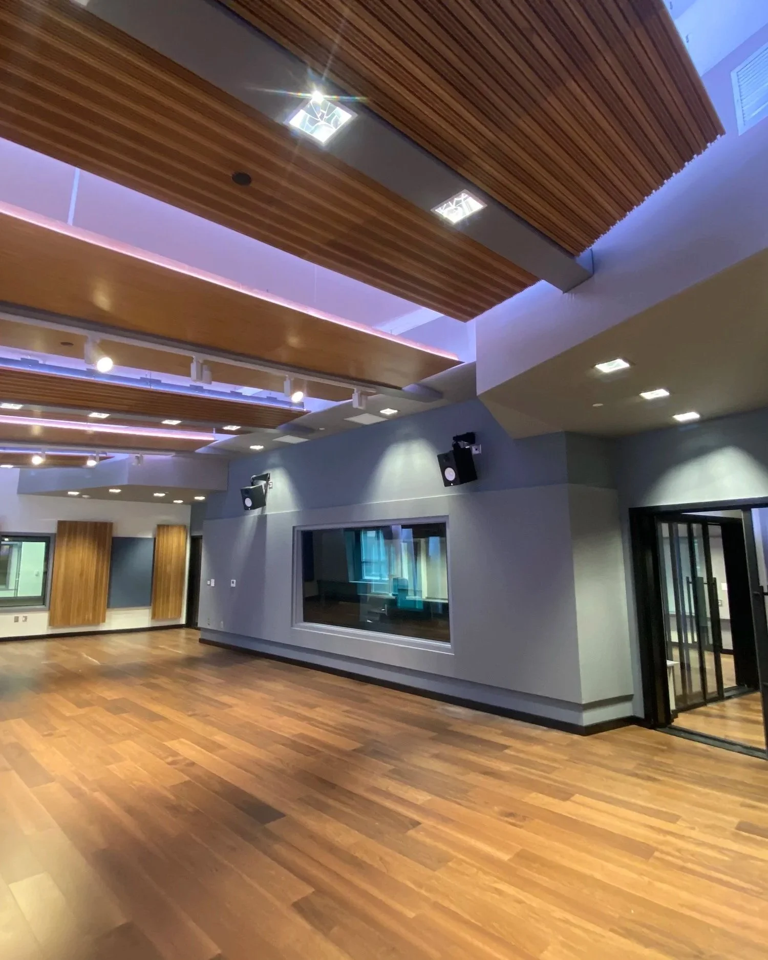 Interior of a modern room with wooden flooring and decorative wooden ceiling panels, speakers on the wall, large window, and glass door.
