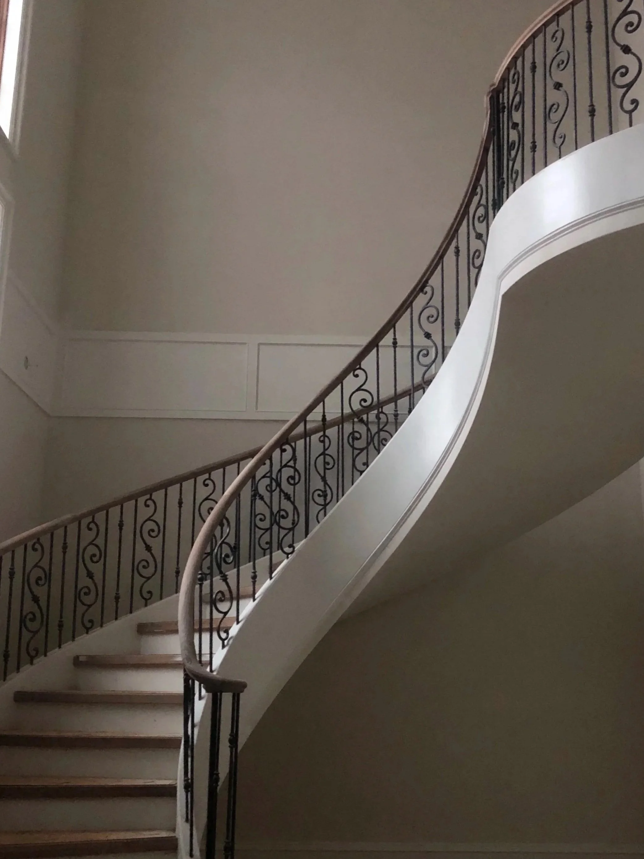 Interior view of a staircase with wooden steps and ornate wrought-iron railing, curved design, ascending from the bottom left to the top right of the image. The staircase is against a beige wall with decorative molding, and natural light is coming from a window on the left side.