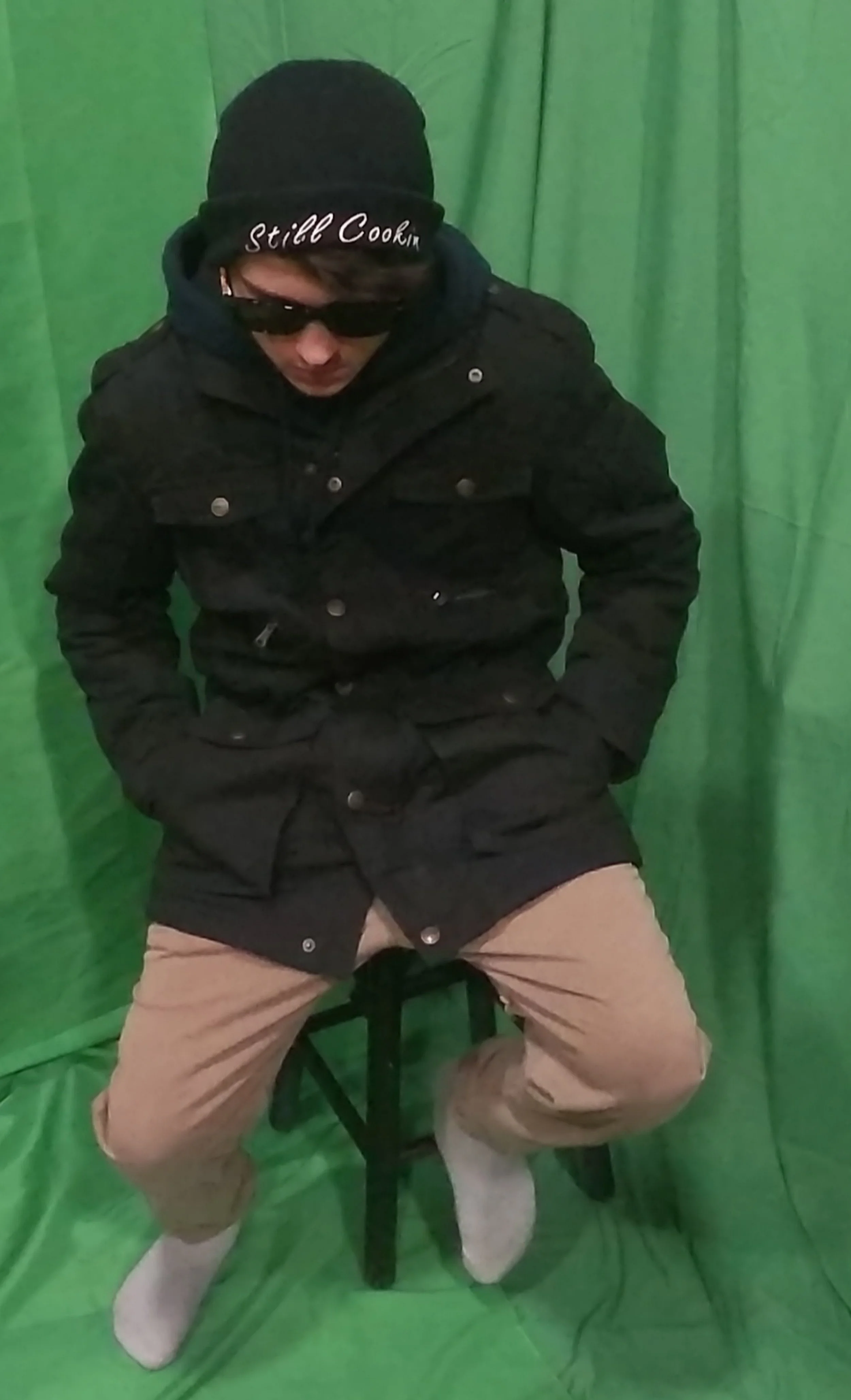 Ciccone sitting on a stool doing A basic pose in front of a green screen, wearing a black peecoat, tan pants, black sunglasses, a black still cookin' winter beanie and looking down while holding their hands in their lap/pockets.