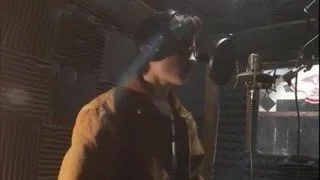 Ciccone posing infront of his microhpone with black glasses & baige jacket while recording vocals in his studio's booth