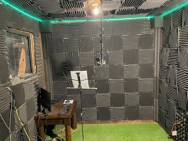 Ciccone's recording studio booth with soundproofing/foam panels on the walls, a computer monitor, portable keyboard & mice on a wooden desk. A window on the left, Green LED strip lighting along the wooden ceiling that matches the fake grass carpeting