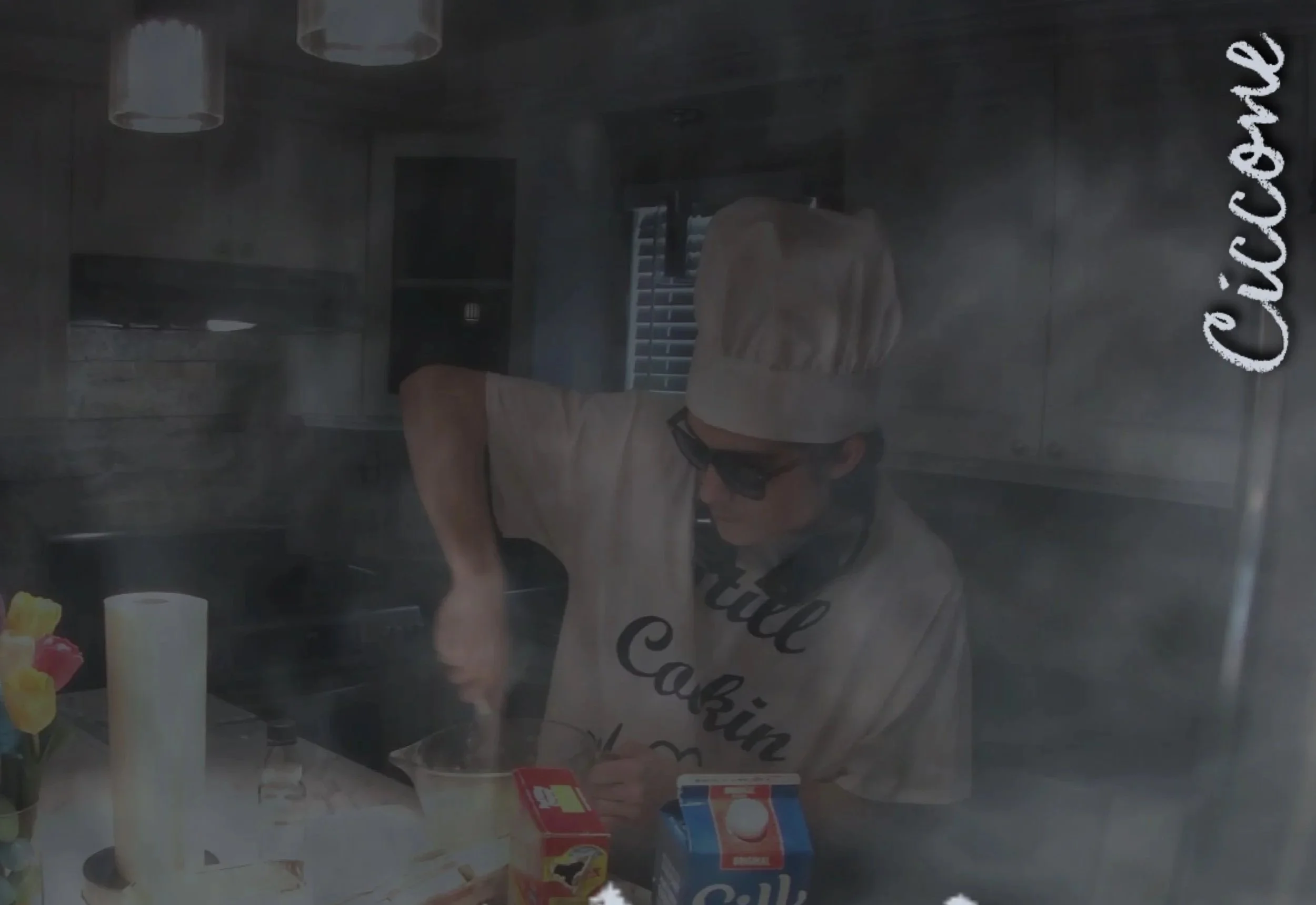 Ciccone wearing A cheff hat, black glasses, and a white still cookin' shirt while whisking/mixing A bowl of ingrediants in A kitchen.