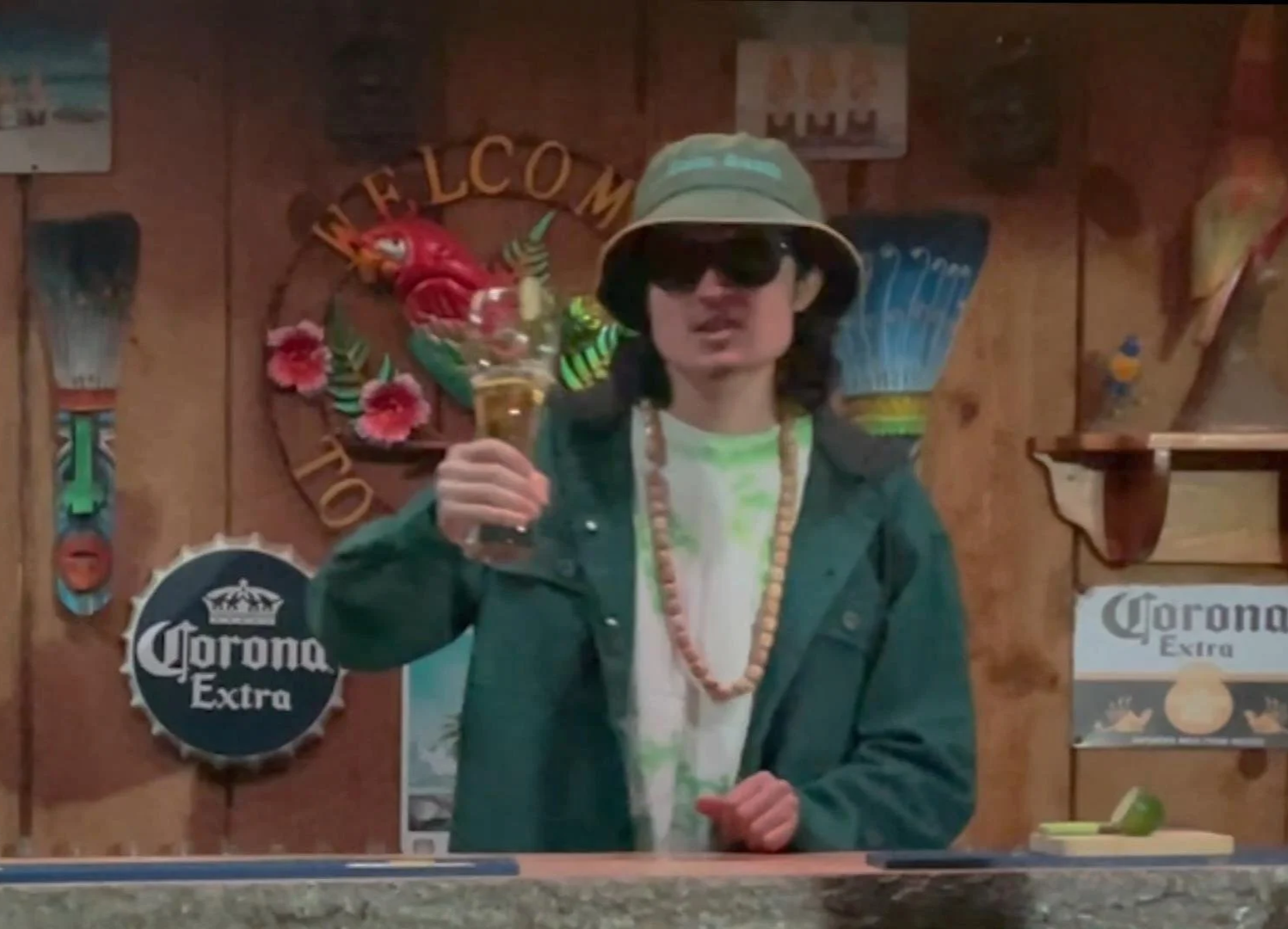 Ciccone wearing A Balm Beach bucket hat, black sunglasses, and a green jacket holding a beer showing A ''cheers'' while smiling towards the camera at his tiki bar decorated with tropical and beer-themed things.
