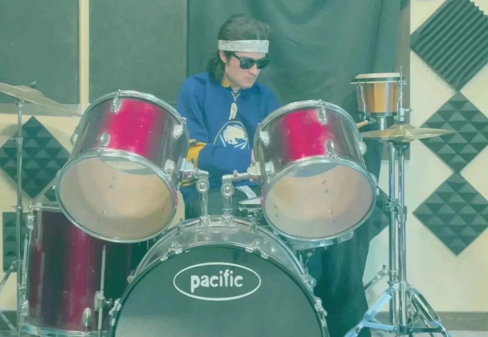 Ciccone playing A drum set in his music studio, wearing black sunglasses, a white bandana, and a bluebuffalo sabers jersey #11