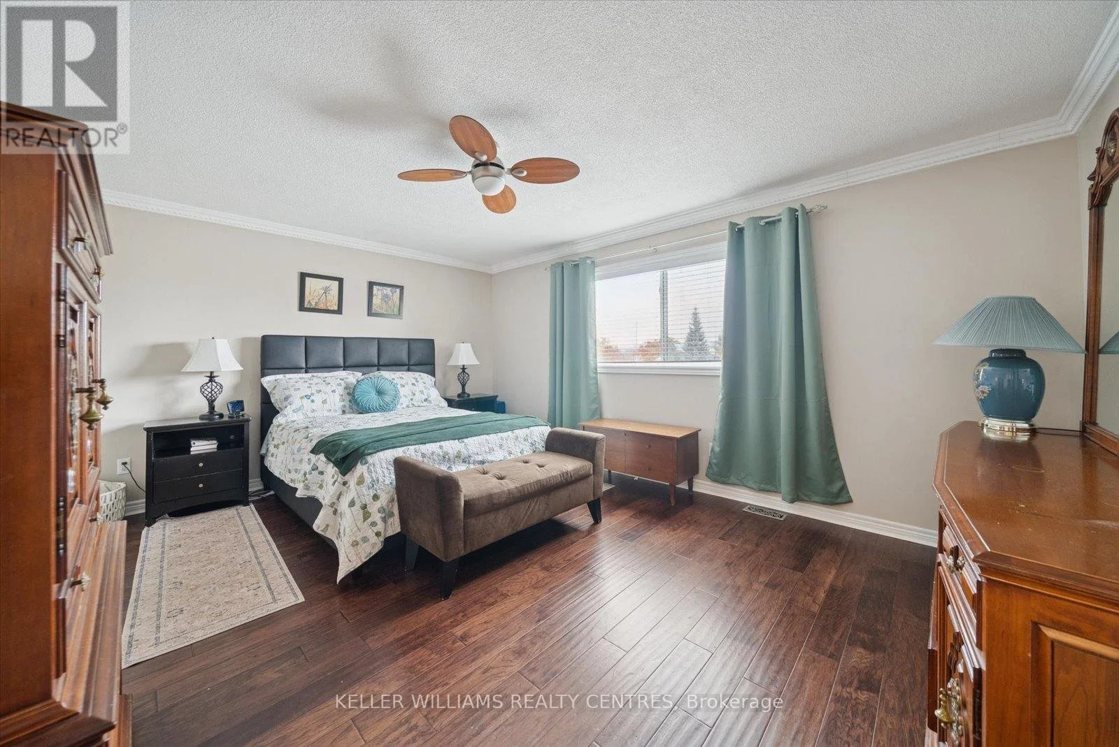 A bedroom with a large bed with a black headboard, two small black nightstands with lamps, a beige bench at the foot of the bed, a window with green curtains, a wooden dresser with a blue lamp, and hardwood flooring.