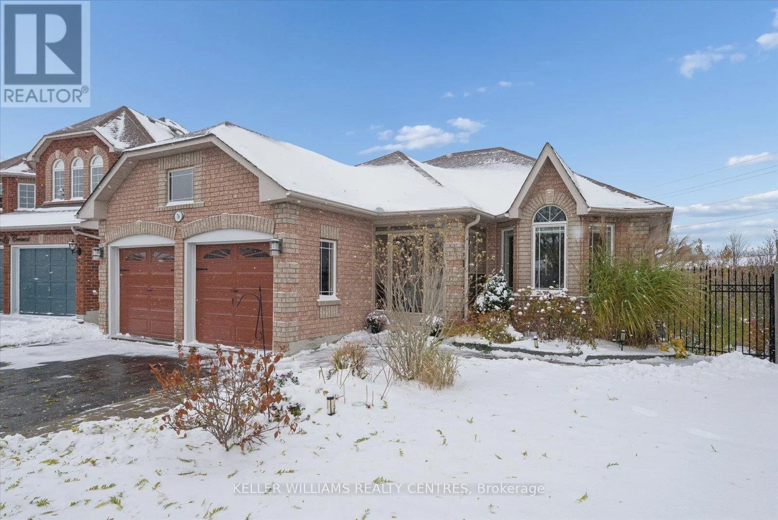 A two-story brick house with a snow-covered roof and yard, brown garage doors, and a front porch with plants and shrubs, in a winter setting.