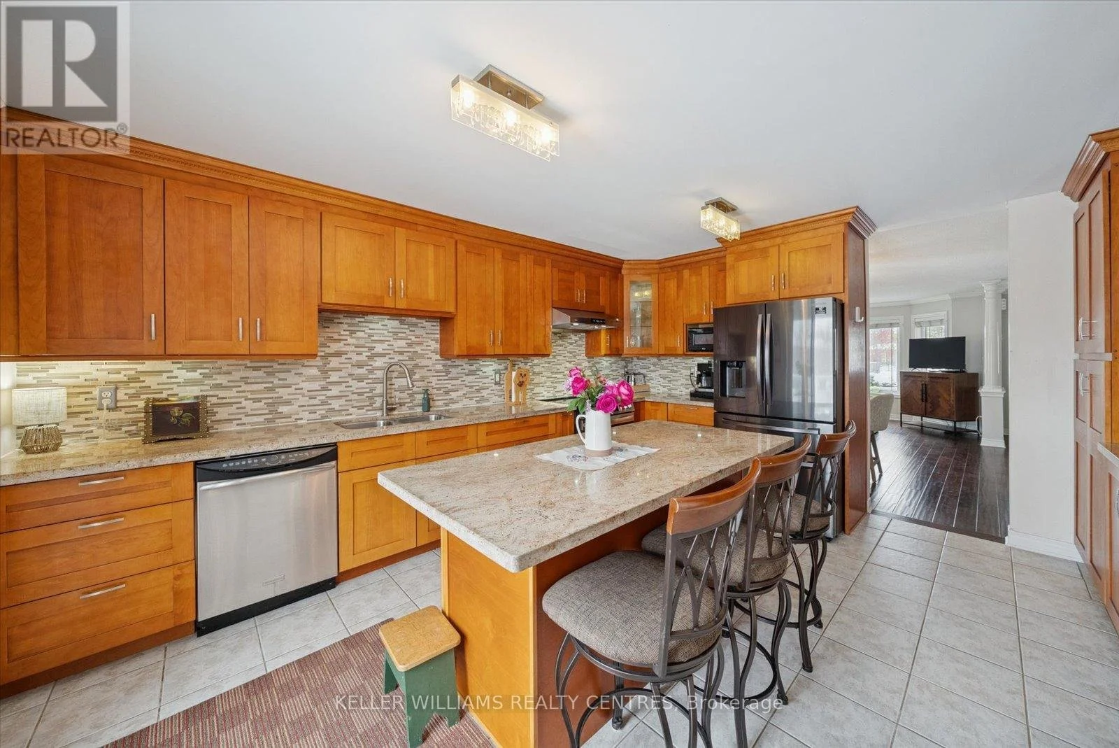 Kitchen with wooden cabinets, granite countertop island, stainless steel appliances including refrigerator and dishwasher, mosaic backsplash, and a dining area with four bar stools. There is a vase with pink flowers on the island and a view into the living room with a TV.