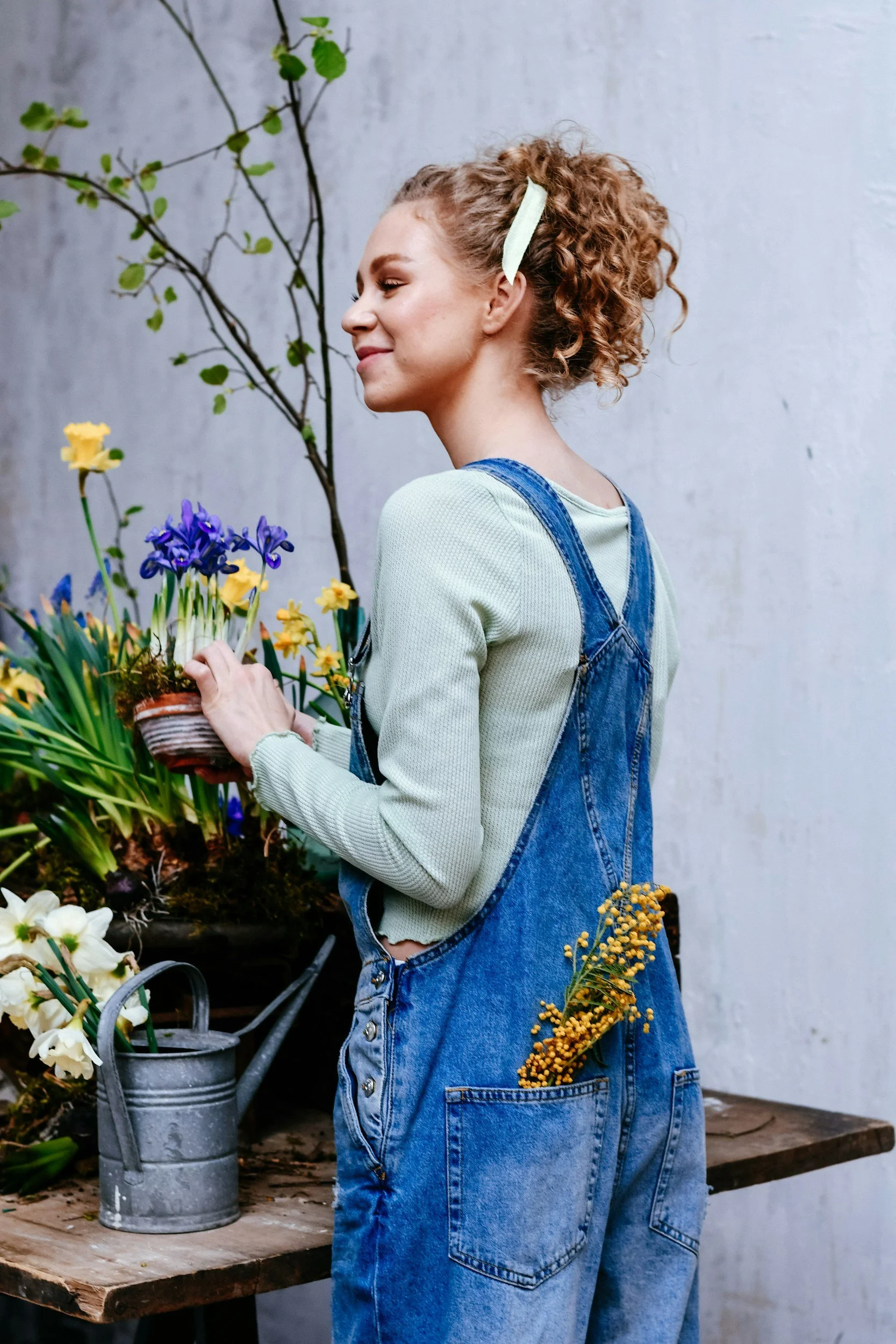 A woman in denim overalls and a light green long-sleeve shirt holding a basket of colorful flowers, standing near a wooden table with gardening tools and a watering can against a gray wall.