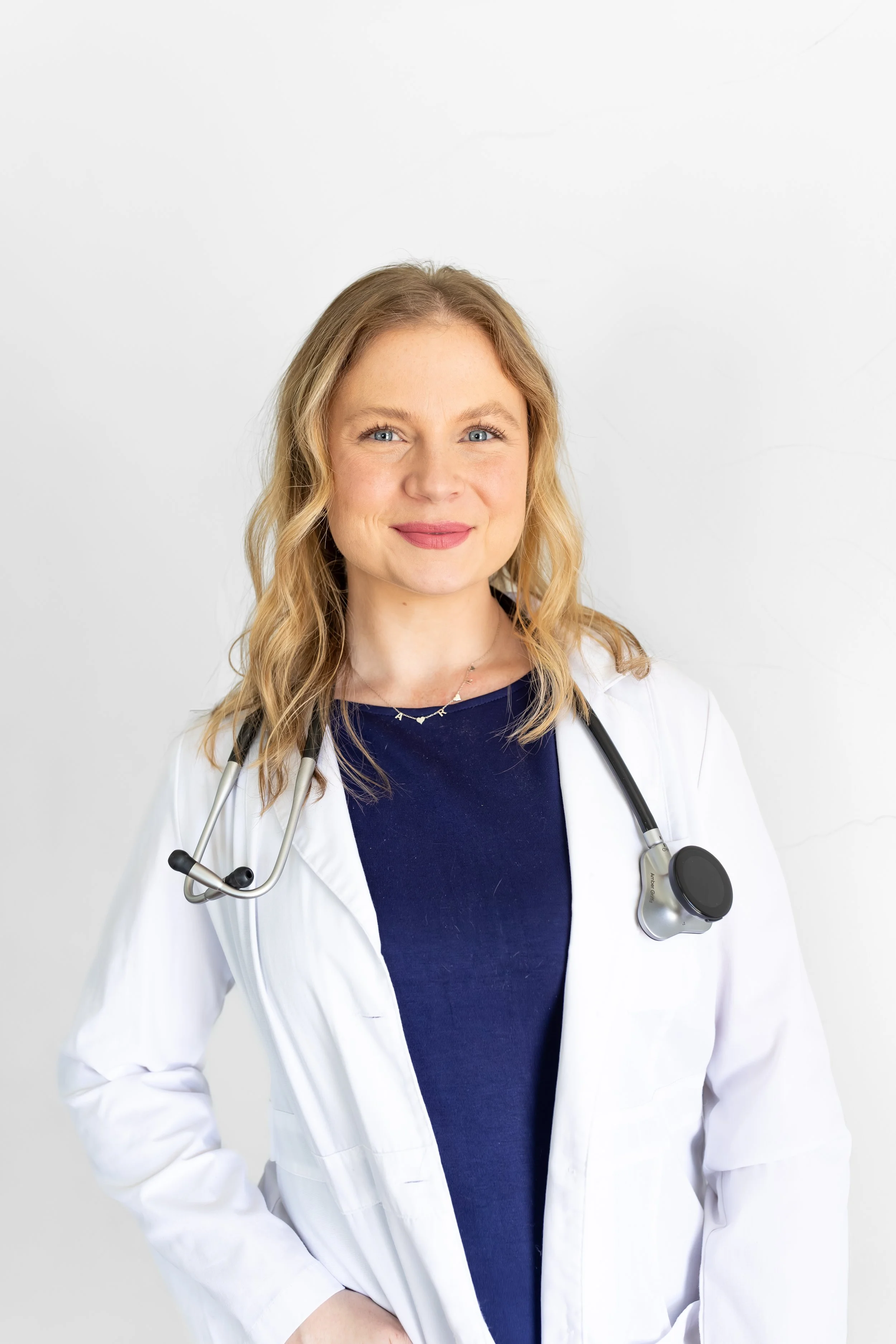 A female doctor with blonde hair and blue eyes, wearing a white lab coat and stethoscope around her neck, standing against a plain white background.