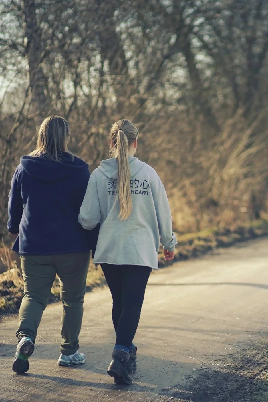 Two women walking together outdoors on a dirt path, surrounded by trees.