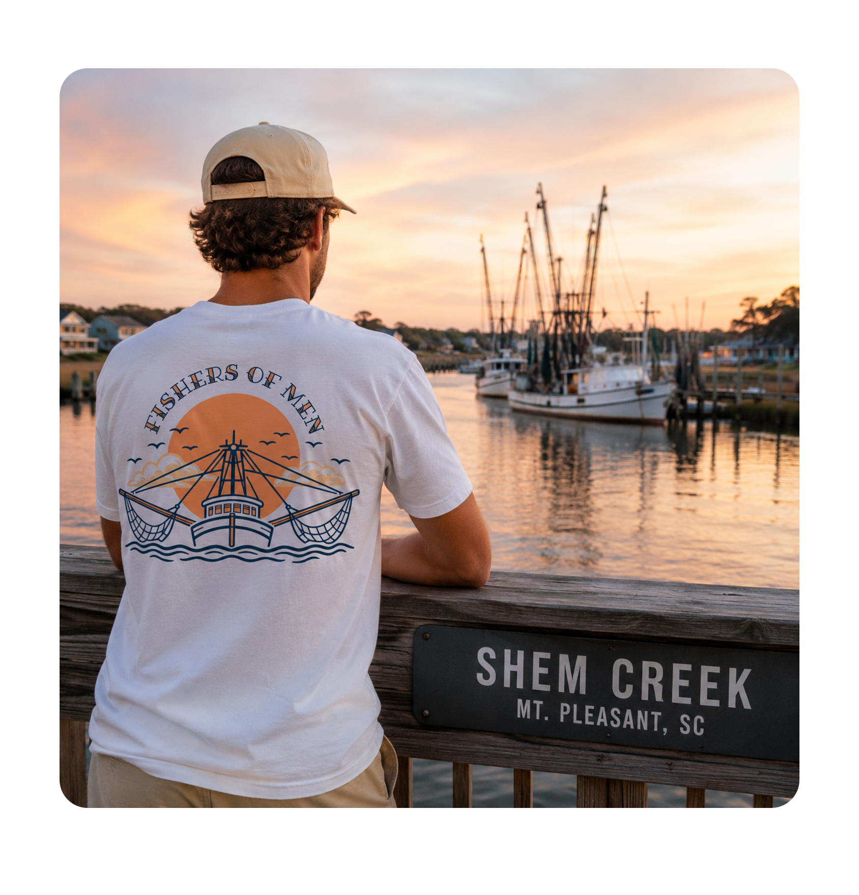 A man with curly hair wearing a beige cap and a white t-shirt with a fishing-themed graphic, standing at a wooden railing overlooking boats on the river at sunset. There is a sign on the railing reading "Shem Creek, Mt. Pleasant, SC."