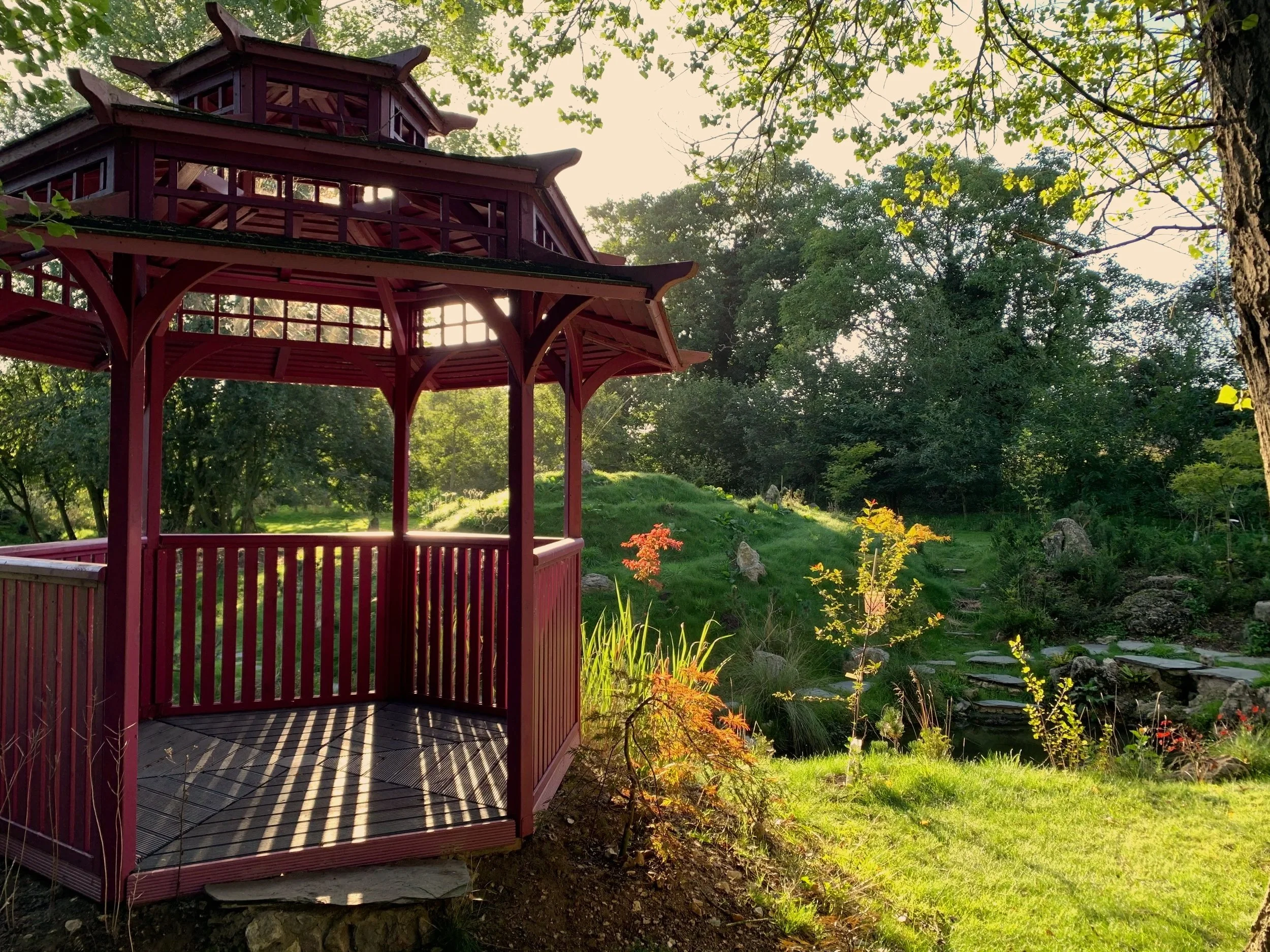 A red Asian-style pagoda with an open lower level in a lush garden with trees, plants, and rocks, during daylight.