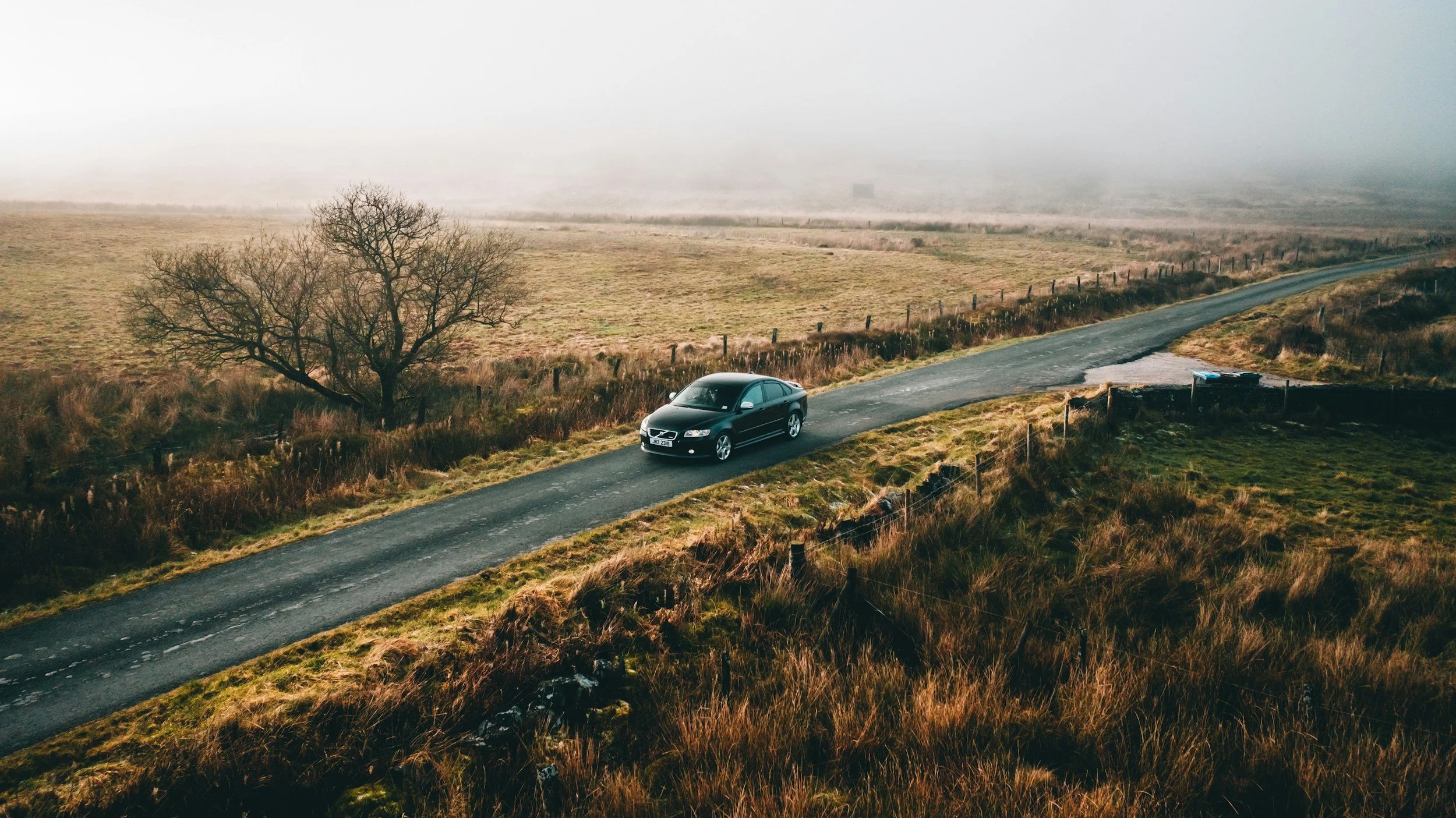 A black sedan car driving on a country road through a foggy, open field with sparse trees and dry grass.
