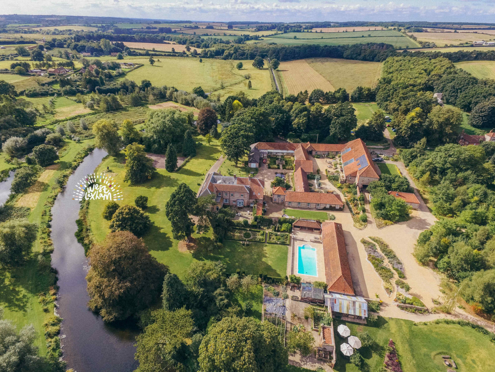 Aerial view of a large countryside estate with multiple buildings, a swimming pool, a river, and lush green fields and trees.