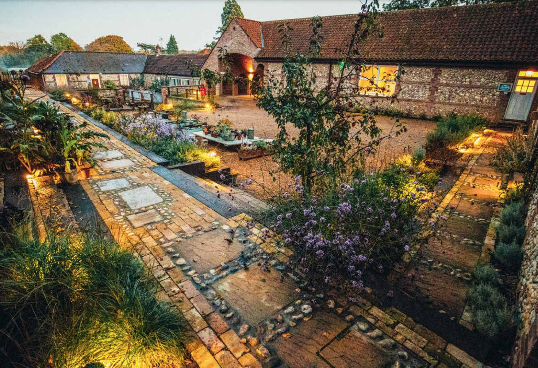 A charming village scene at dusk with cobblestone paths, flowers, and old brick buildings illuminated by warm outdoor lights.