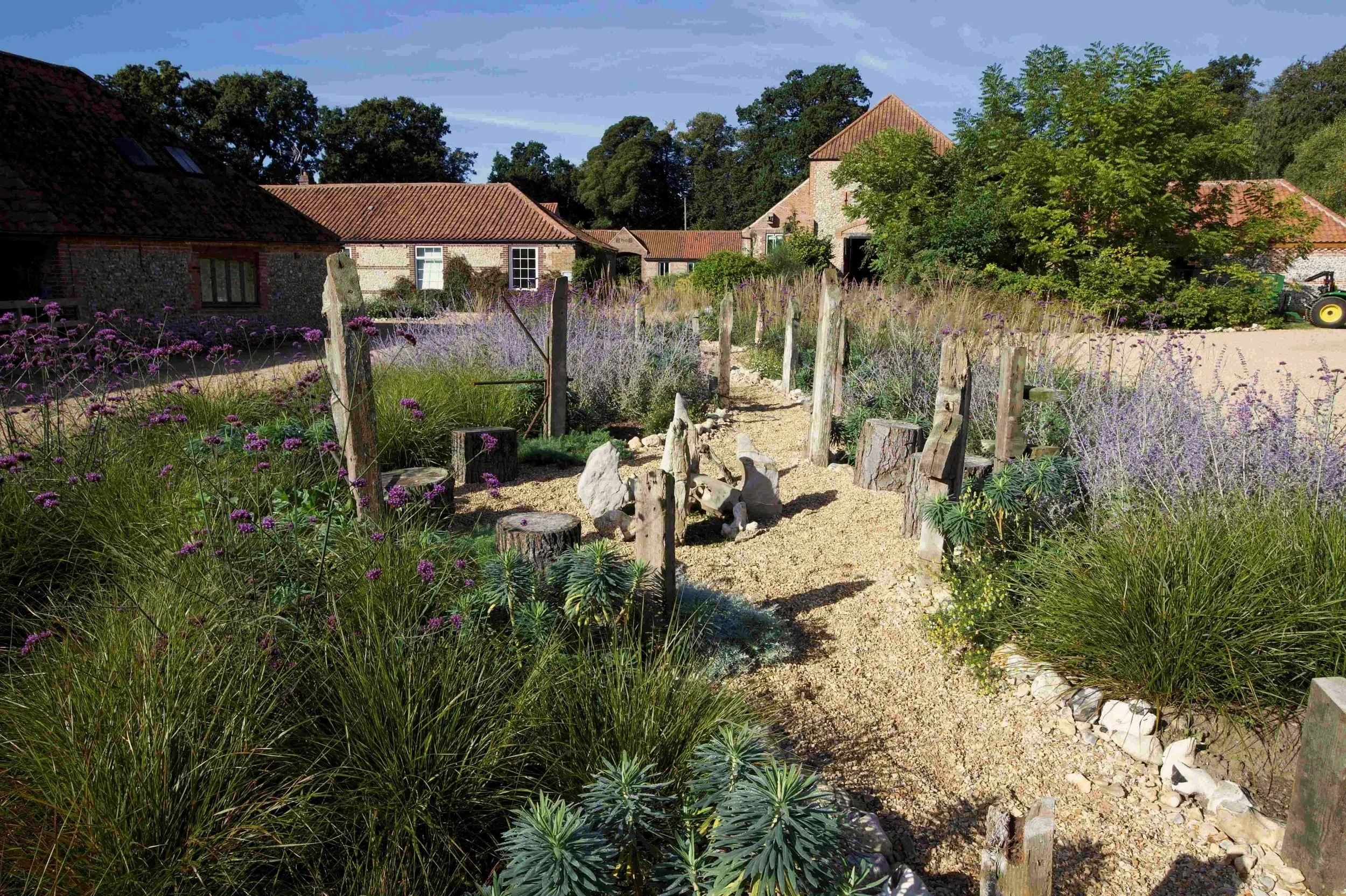 A rustic farmyard scene with old brick buildings, a garden, and a dirt pathway. There is greenery, purple flowers, and a tractor in the background under a clear blue sky.