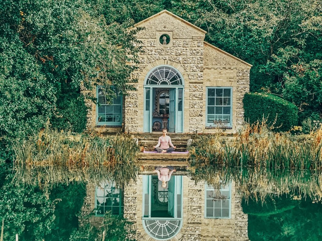 A woman practicing yoga in a seated meditation pose on a mat in front of a stone house by a pond, surrounded by lush greenery, with her reflection visible in the water.