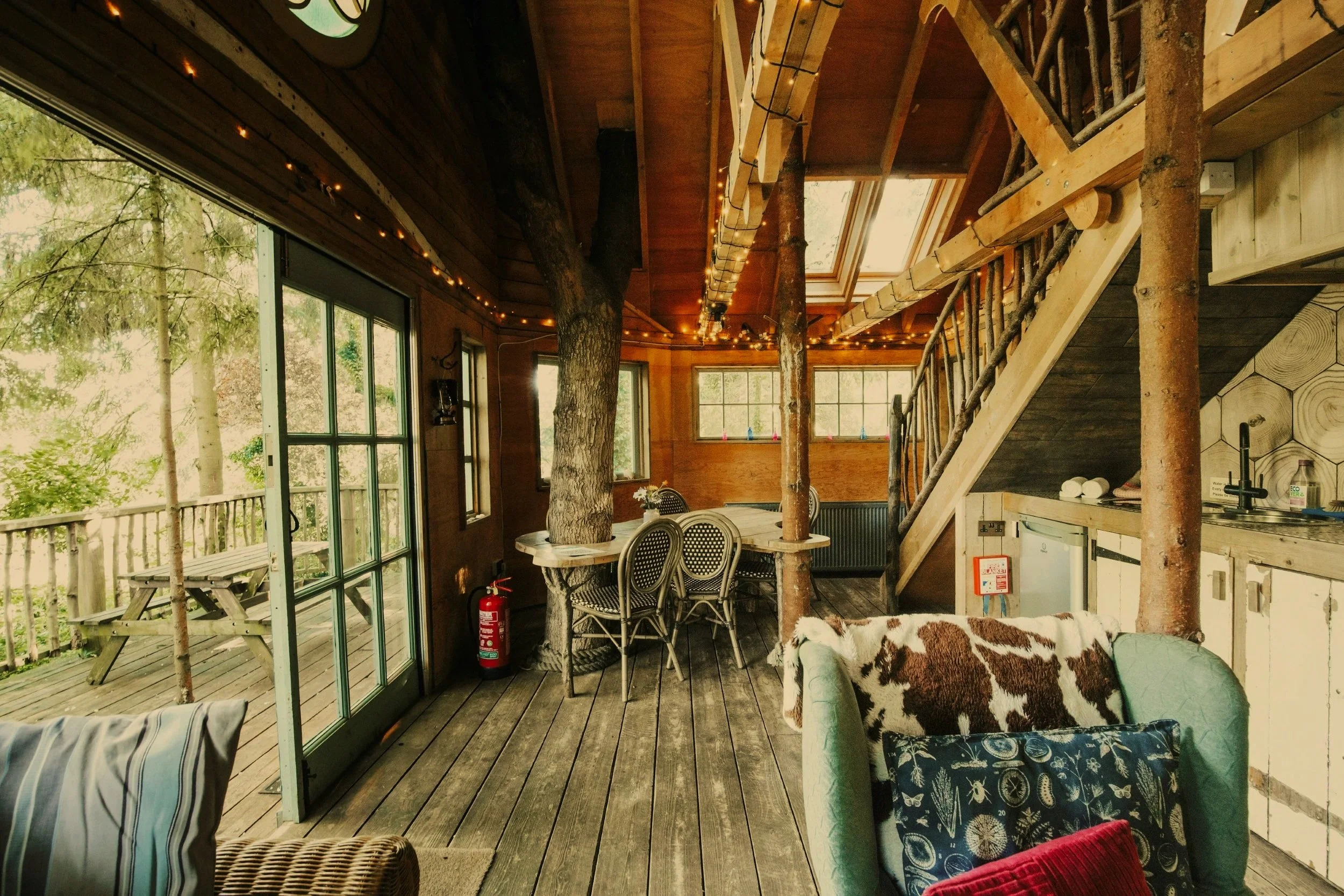 Interior of a rustic cabin with wooden walls and ceiling, large windows revealing a forest outside, and a staircase made of logs leading upstairs. There is a round dining table with chairs, a couch with patterned pillows, and string lights hanging along the ceiling. A fire extinguisher is on the floor near the wall.