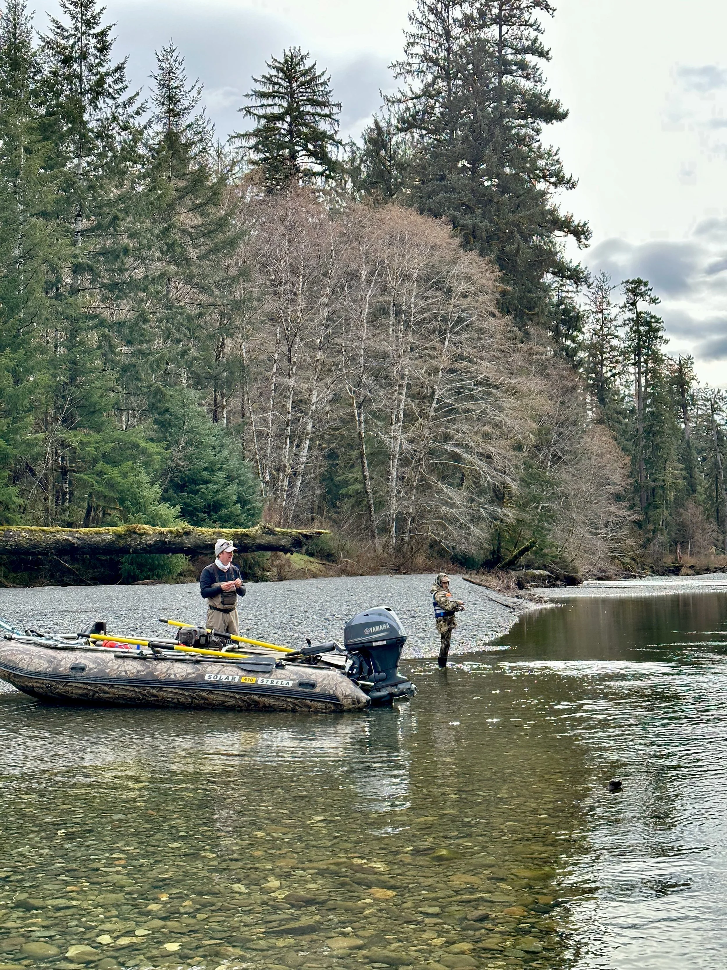 Two men stand by a boat in a river with a forested hillside in the background, one is fishing while the other is holding a fishing rod; the boat has fishing gear and an outboard motor.
