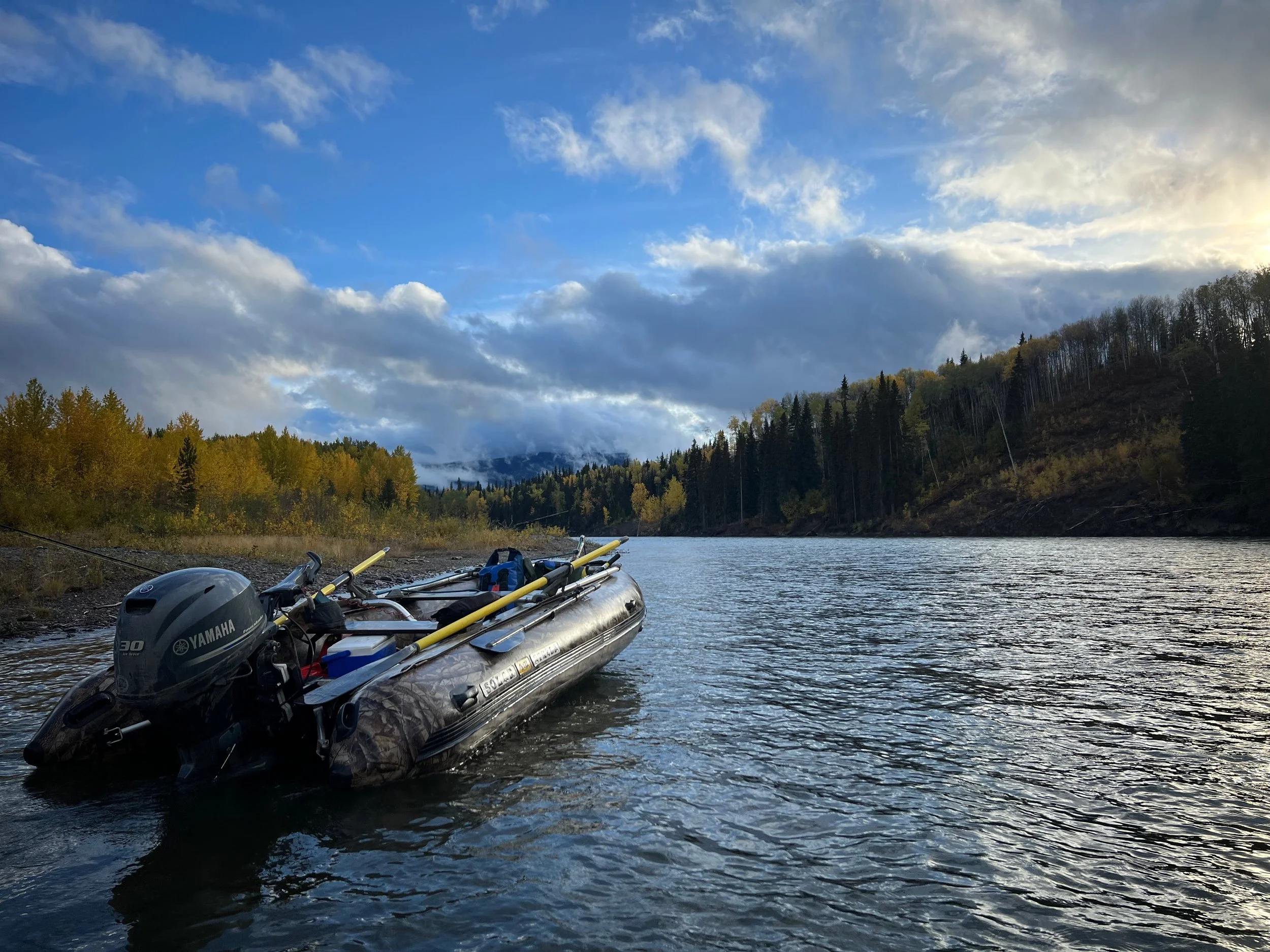 A river with a black inflatable boat containing a Yamaha outboard motor and other equipment, surrounded by forested hills under a partly cloudy sky.