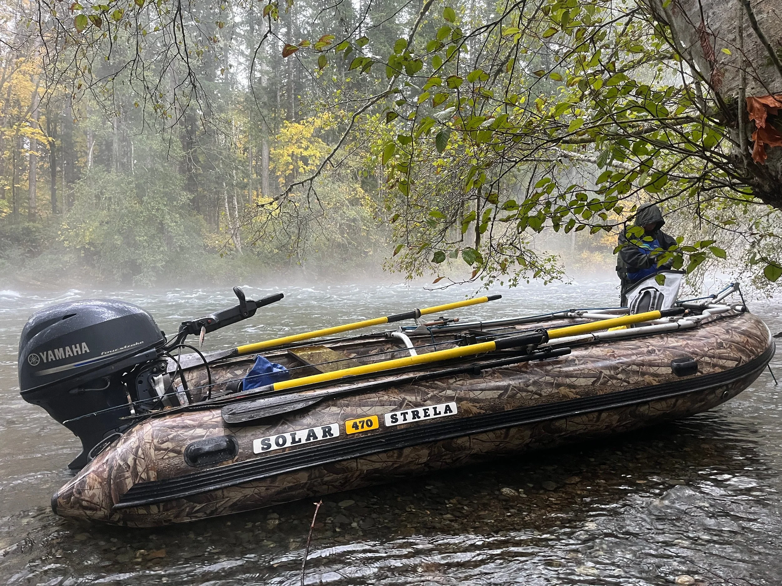 A fishing boat with a Yamaha outboard motor, situated in a river with trees and mist in the background, and a person wearing a hat and jacket on the boat.