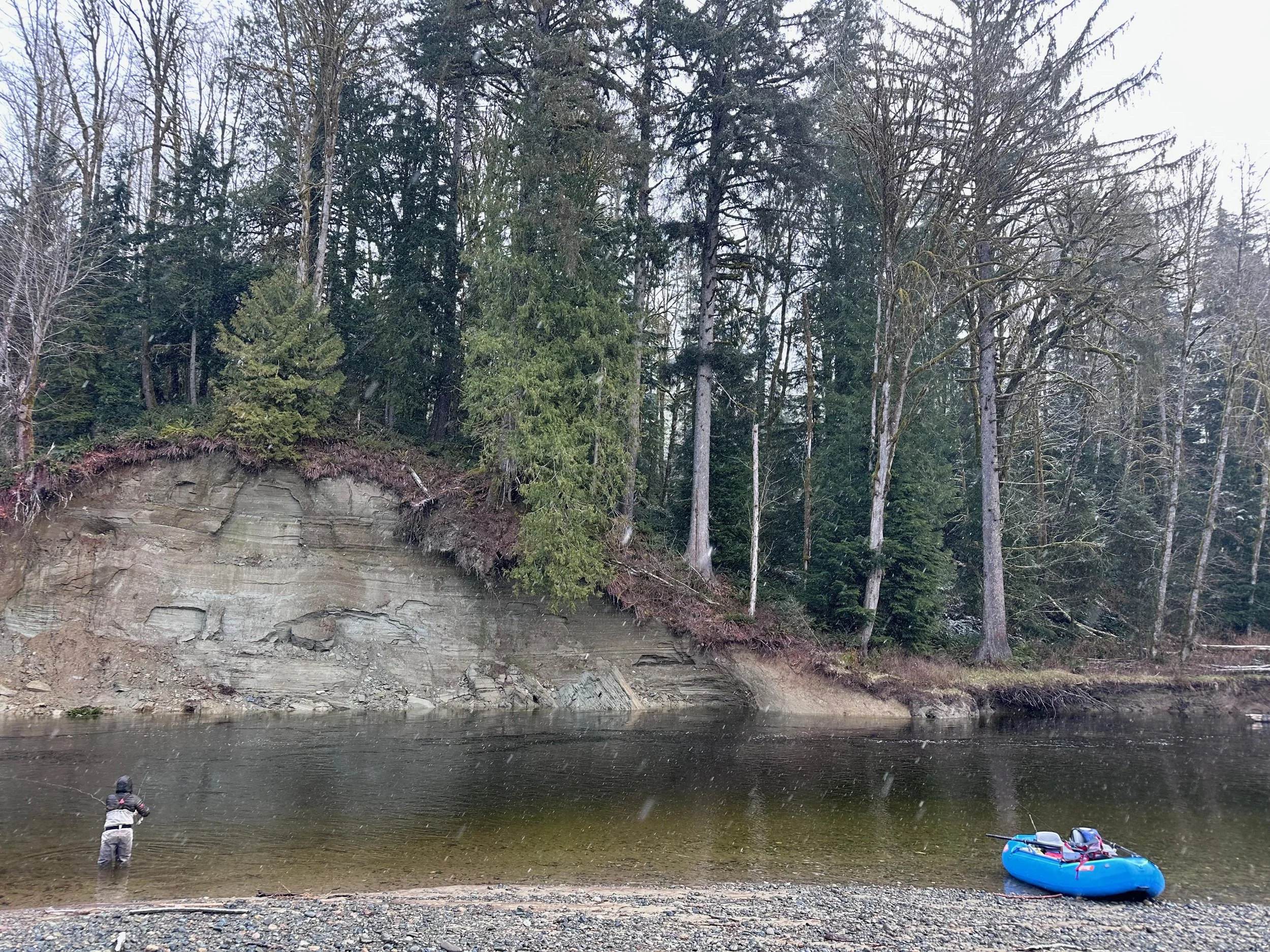 A person fishing in a river with a fishing rod, surrounded by trees in a forested area. A blue inflatable boat is on the riverbank nearby.