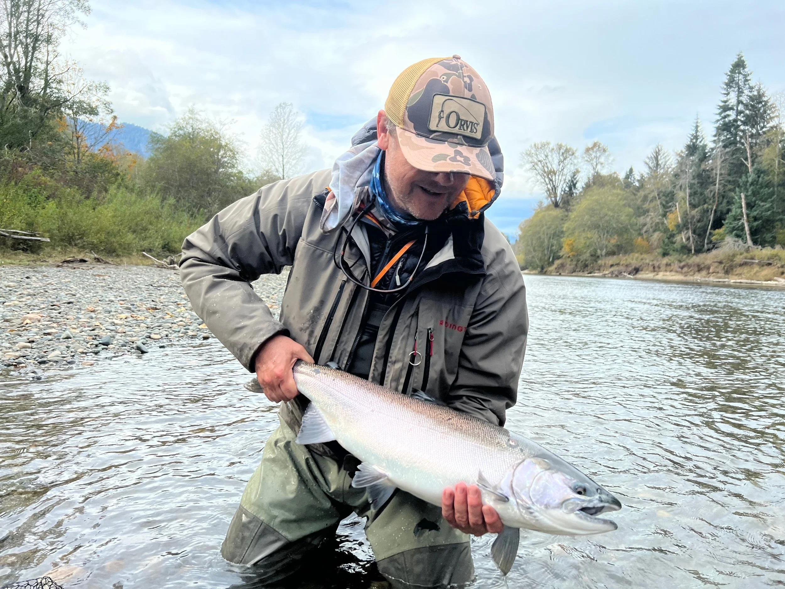 A man in outdoor clothing stands in a river, holding a large coho with both hands. The background features trees with autumn foliage and mountains under a cloudy sky.