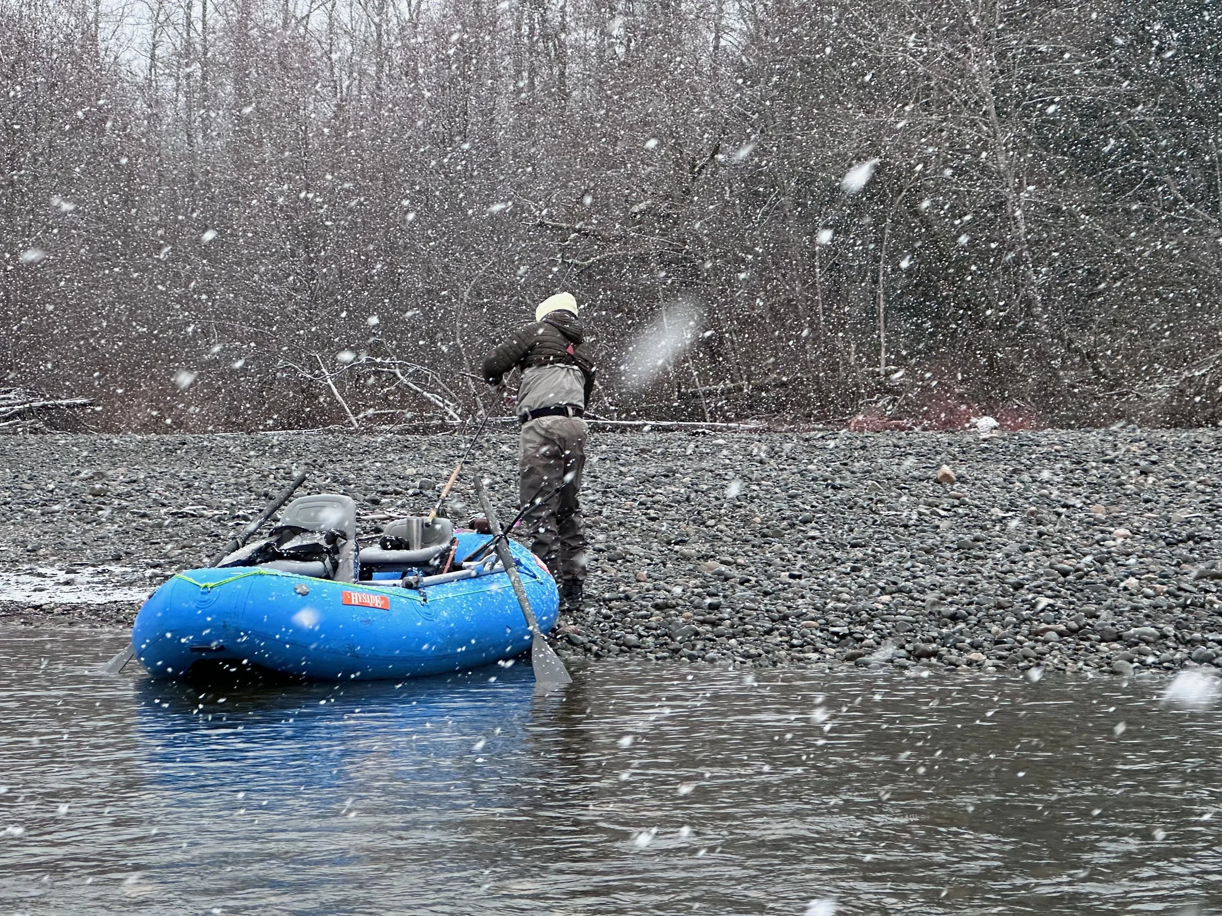 A person in outdoor gear stands near a blue inflatable boat on a rocky riverbank during a snowstorm, surrounded by falling snow and leafless trees.