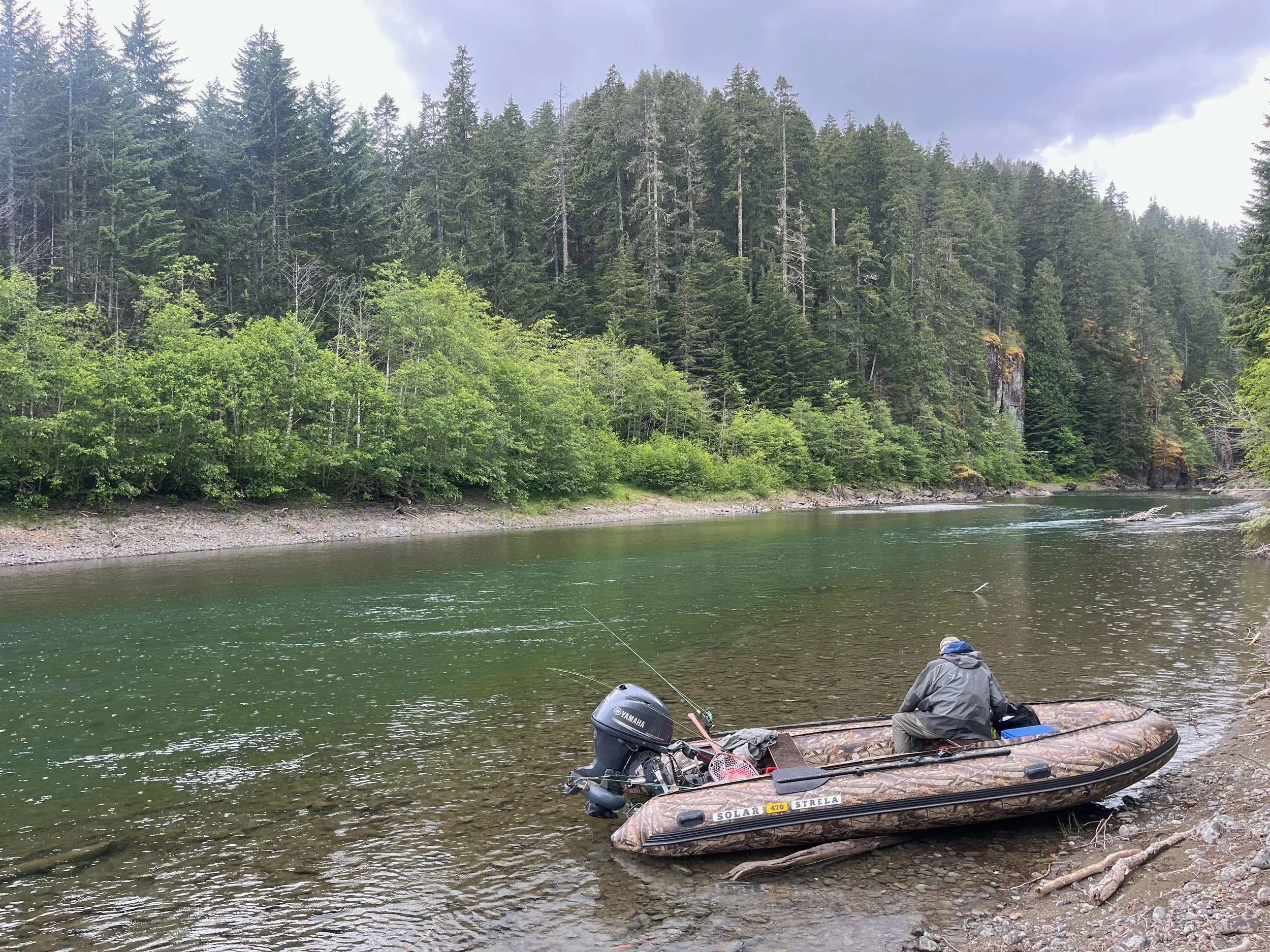 A person sitting on a boat docked at the shore of a river, with fishing gear nearby, in a lush forest with tall trees and a cloudy sky.