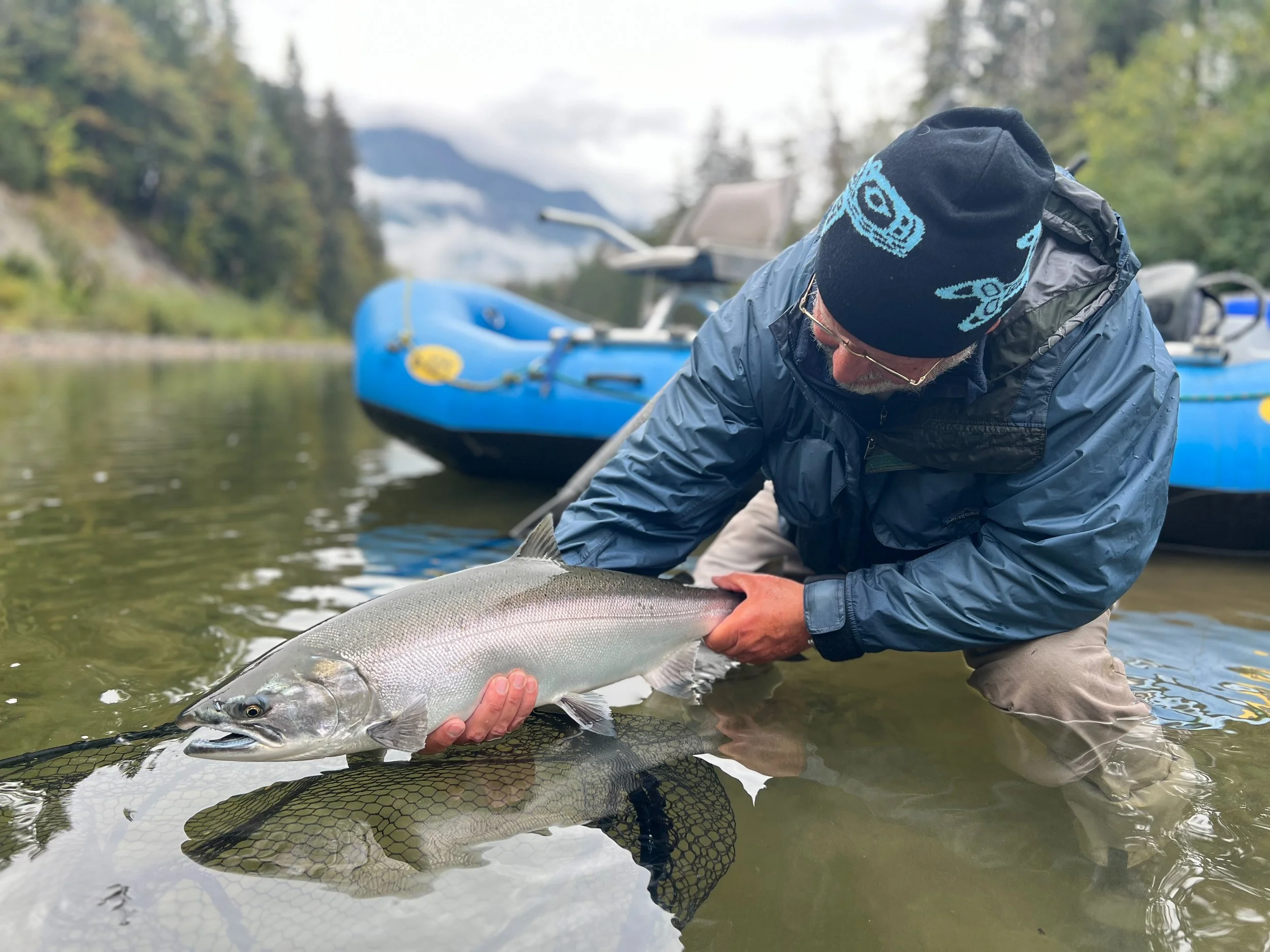 A person wearing outdoor clothing and a black knit cap was catching a large fish in a river, with a blue boat and lush green trees in the background.