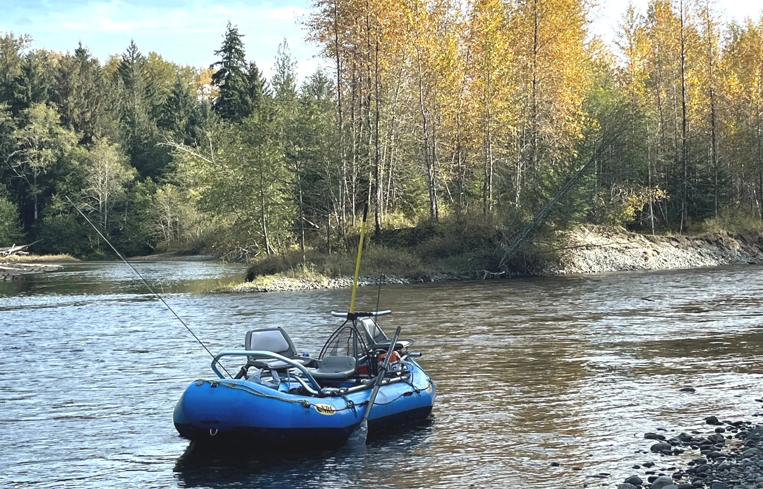 A blue inflatable boat with a fishing rod on a river with a rocky shore, background shows trees with fall foliage and clear sky.