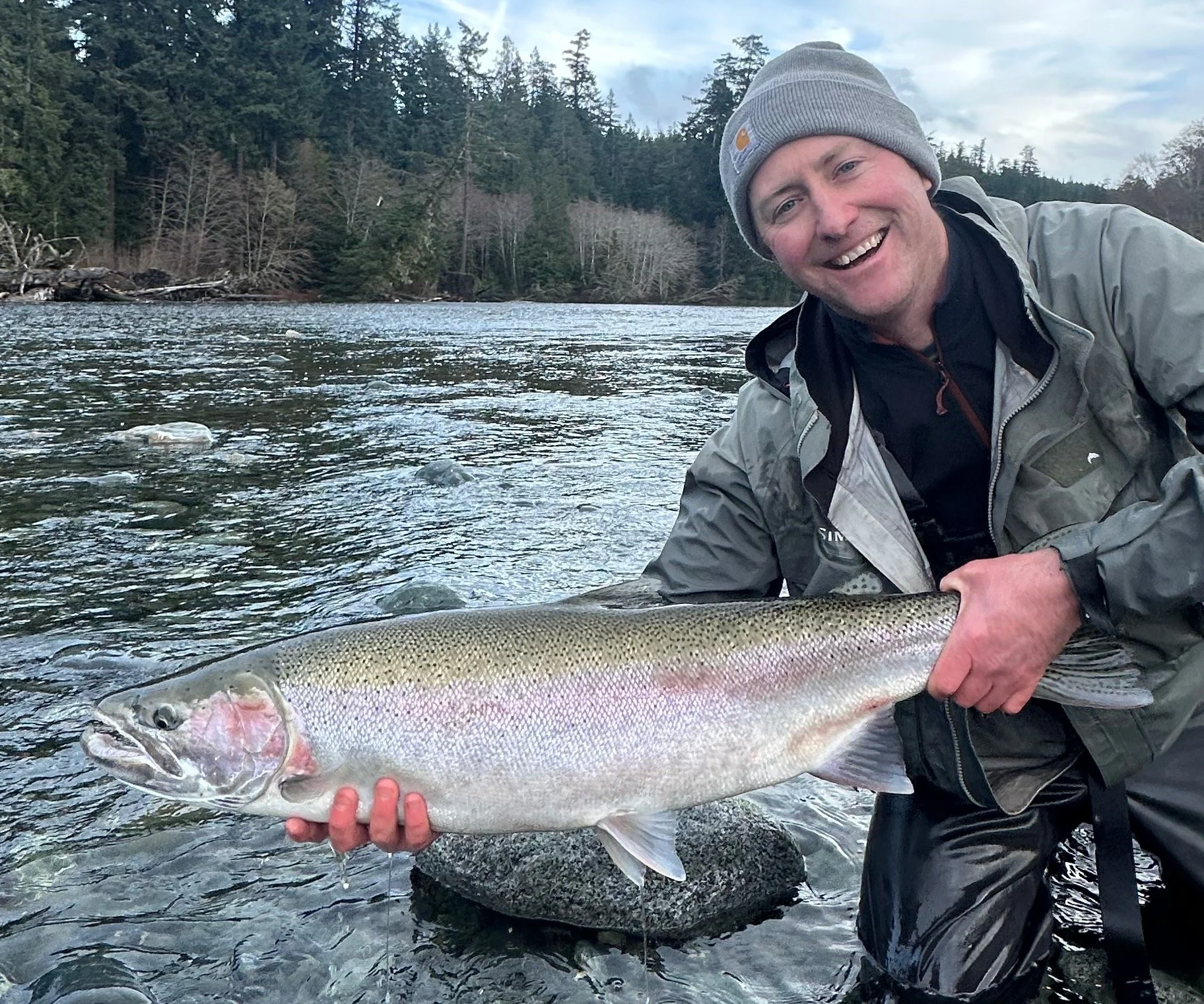 A man in outdoor clothing and a gray beanie hat smiling while holding a large fish he caught in a river with a wooded shoreline in the background.