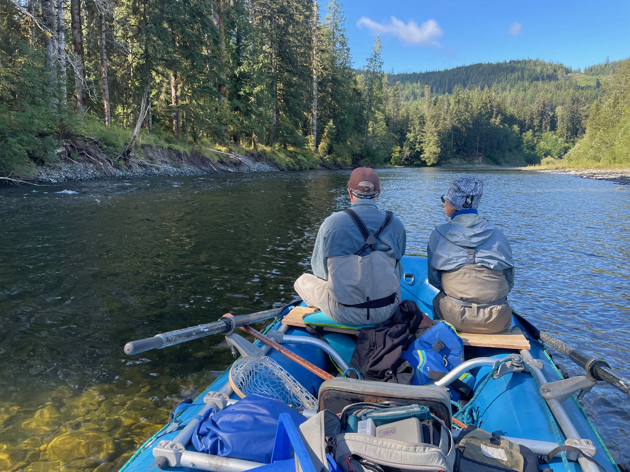 Two people sitting in a blue raft on a river, surrounded by a dense forest and mountains in the distance, under a mostly clear sky.
