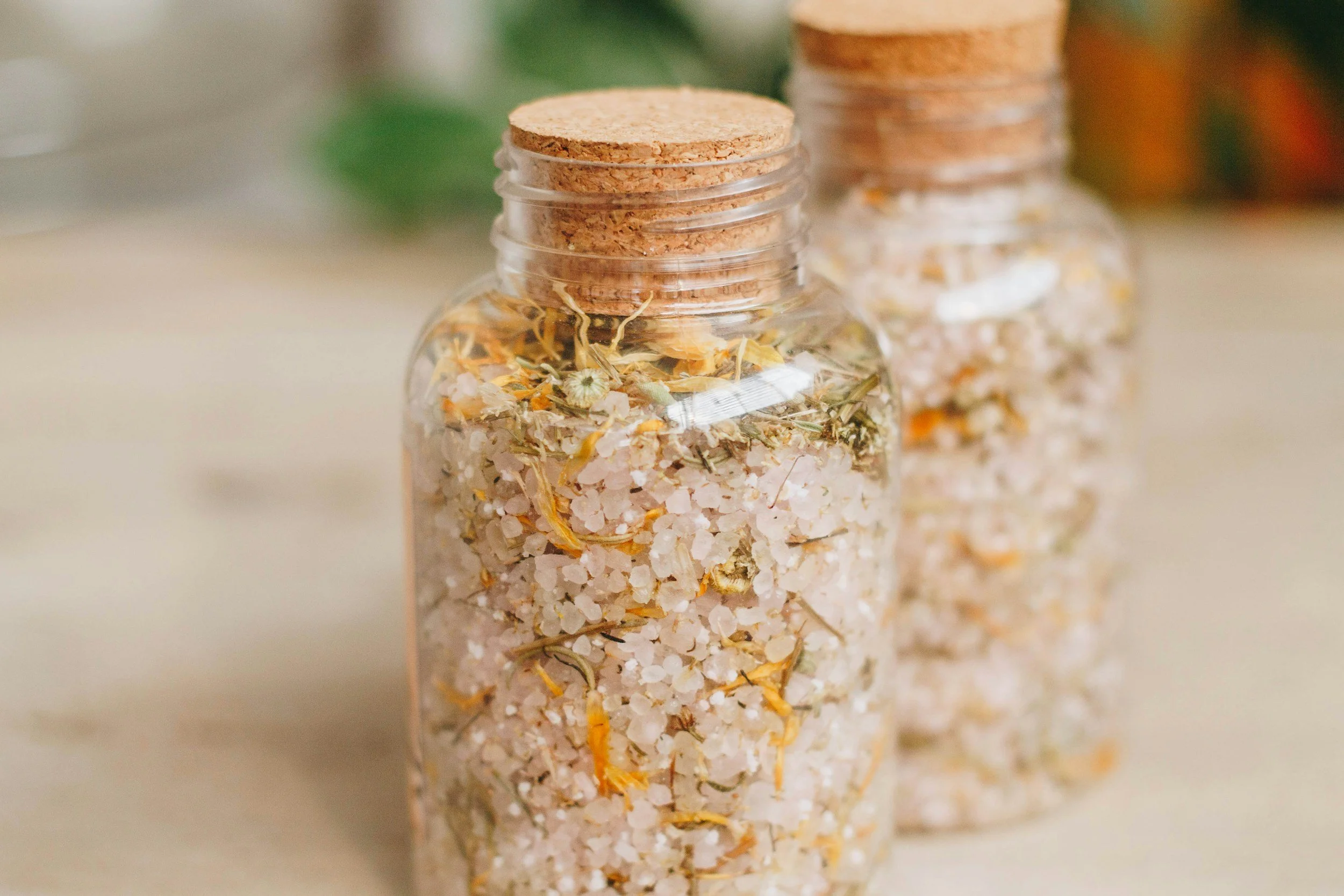 Three glass jars with cork stoppers filled with bath salts and dried herbs.