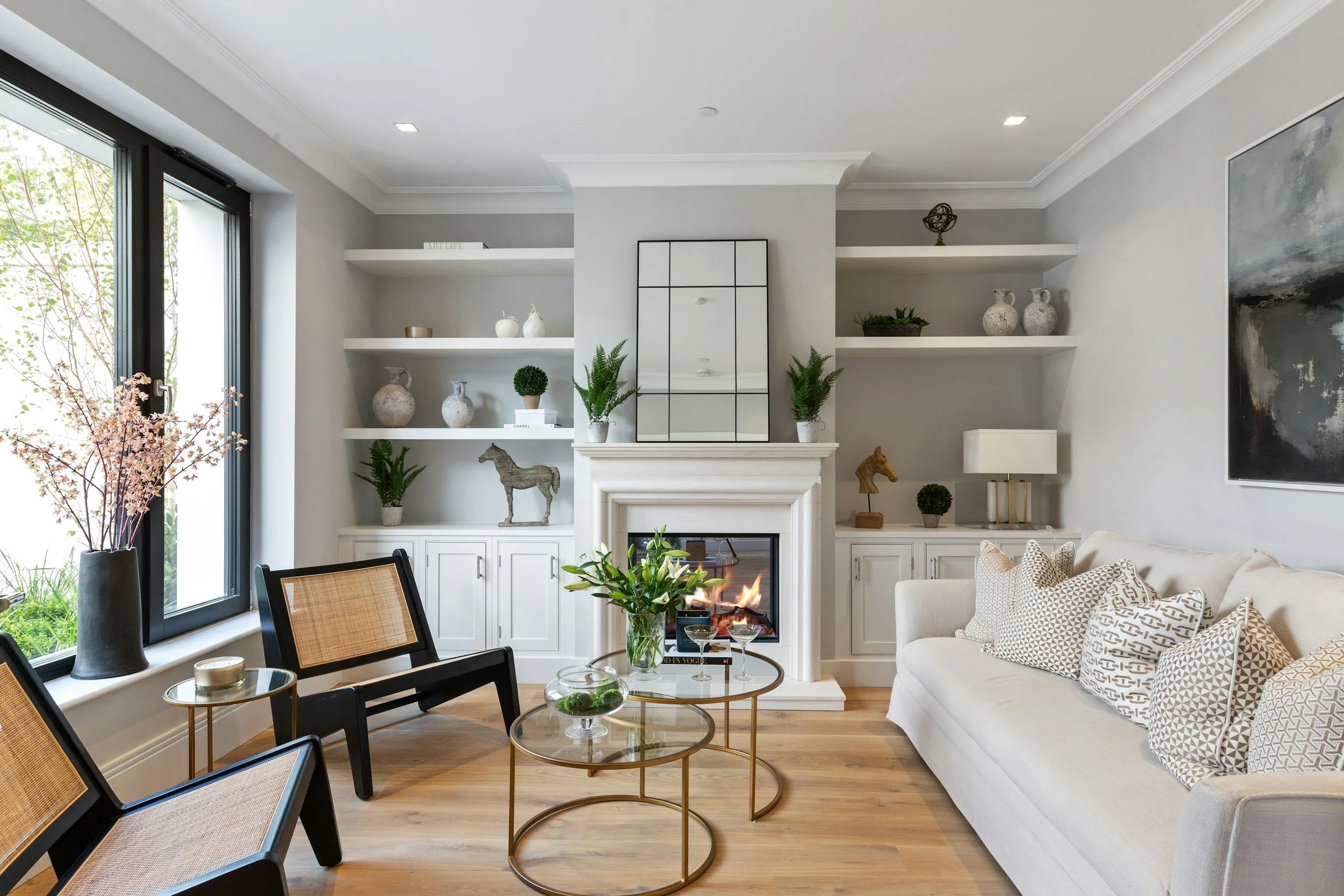 A modern living room with a fireplace, white built-in shelves decorated with vases and plants, a large window with black frames, and a white sofa with patterned cushions. There are round glass coffee tables with decorative items, and a dining area with black chairs and a vase of flowers.
