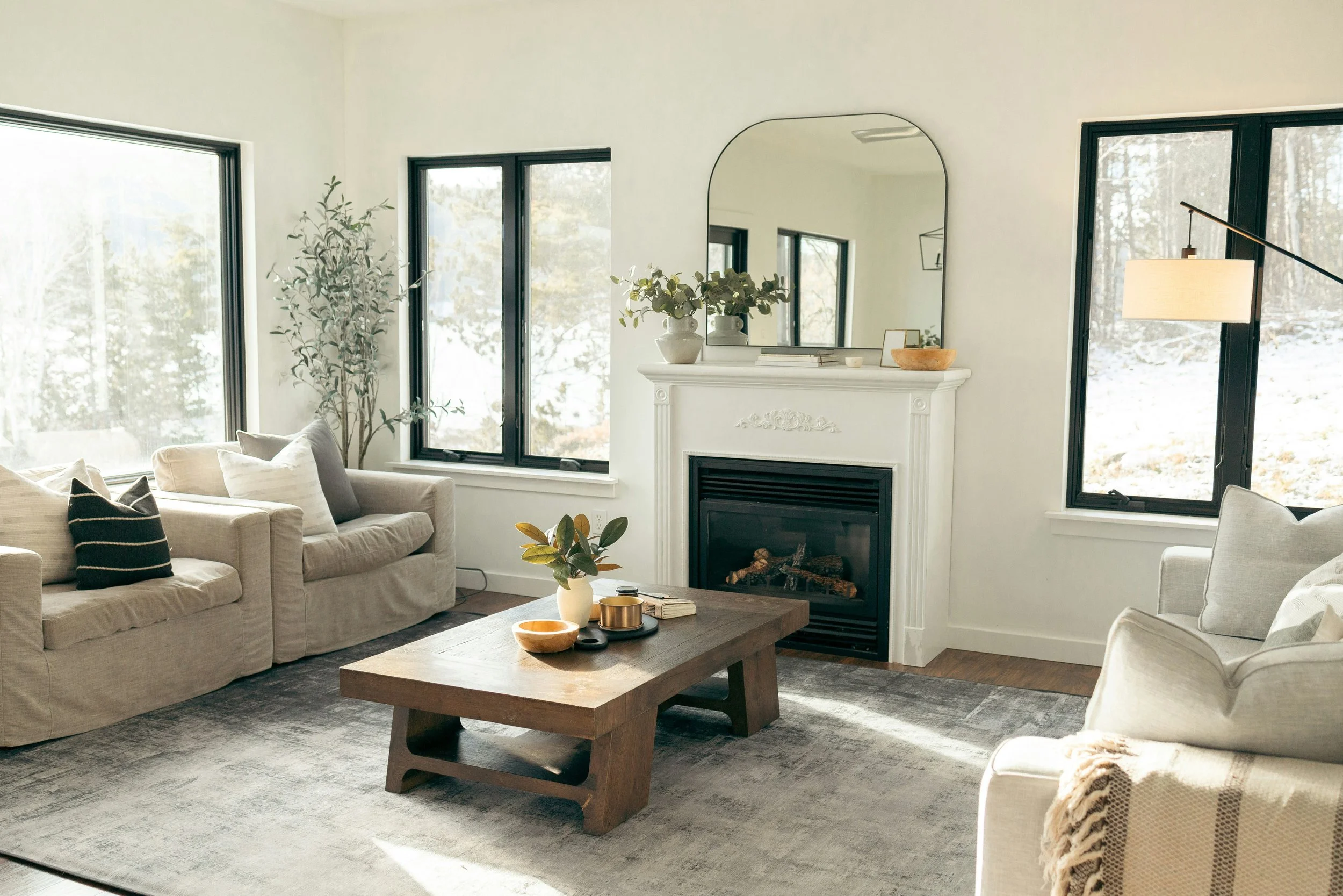 Bright living room with beige sofas, a wooden coffee table, fireplace, large windows showing snowy outdoor landscape, plants, mirror, and modern decor.
